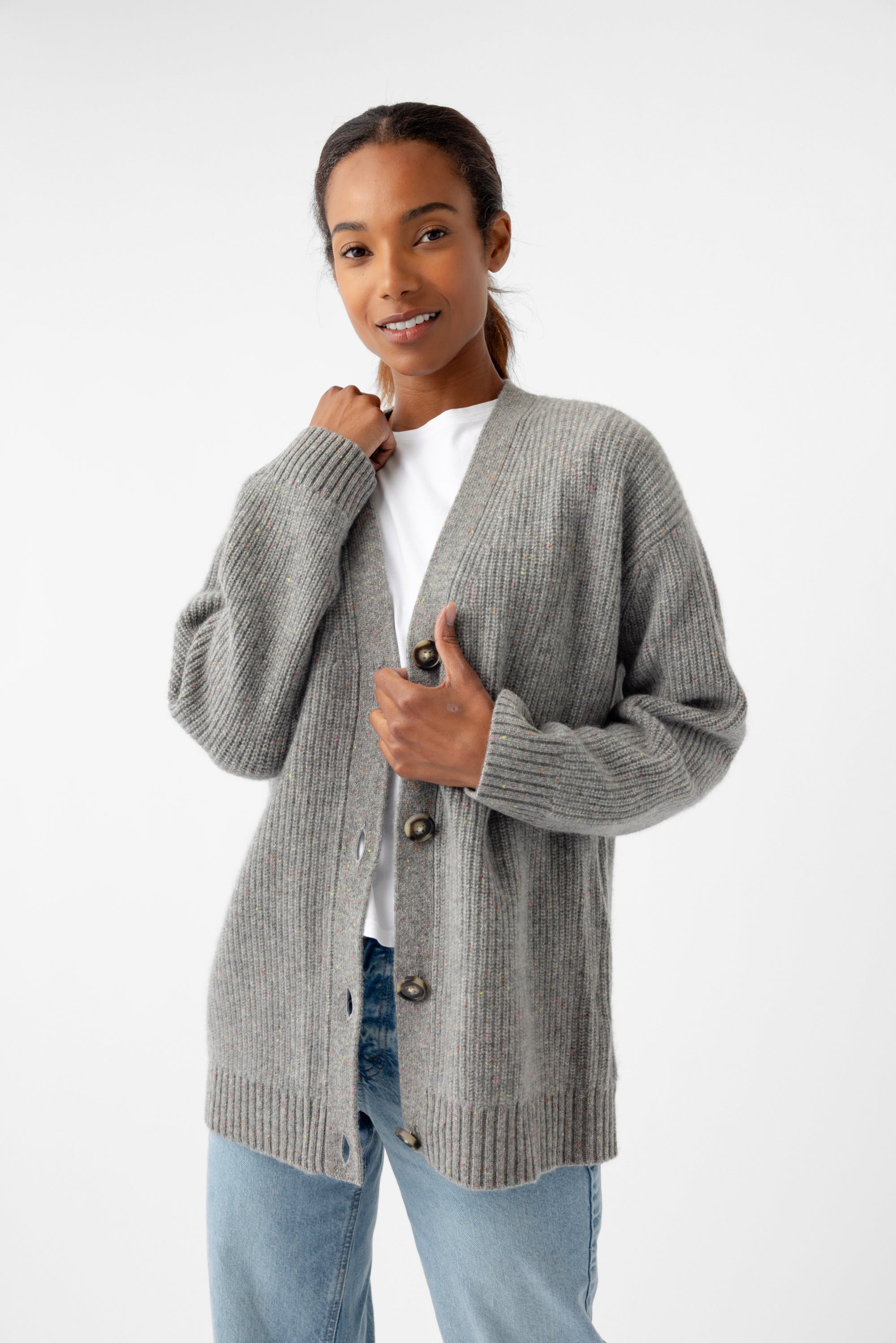 A person is styled in an Oversized Sunday Cashmere Cardigan from Cozy Earth, layered over a white shirt and blue jeans. Standing against a plain white background, they are slightly smiling with one hand gently adjusting the cardigan. 