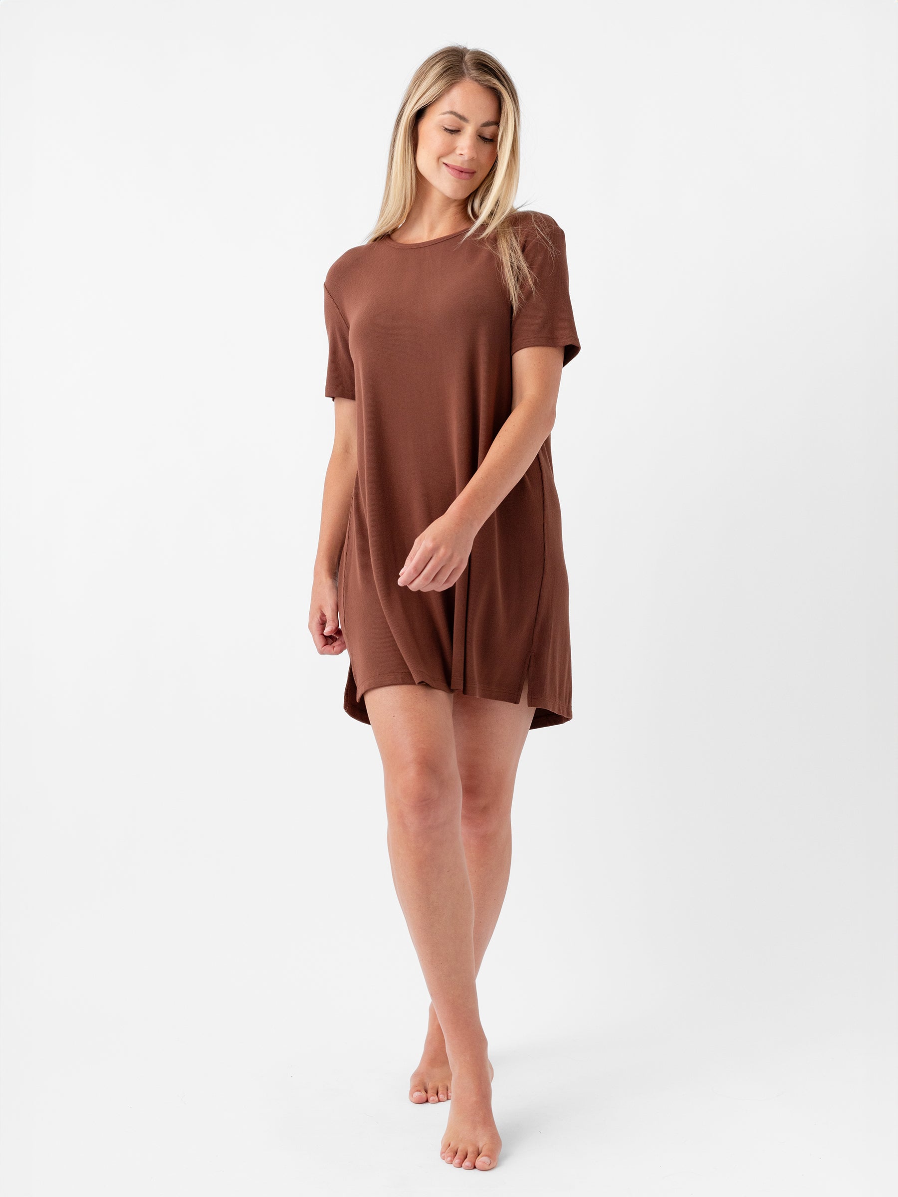 A woman with long blonde hair stands barefoot against a white background, wearing the Cozy Earth Women's Bamboo Rib-Knit Boyfriend Sleep Dress. She looks down, holding the hem of the loose-fitting, short-sleeved brown dress gently with one hand. |Color:Spice