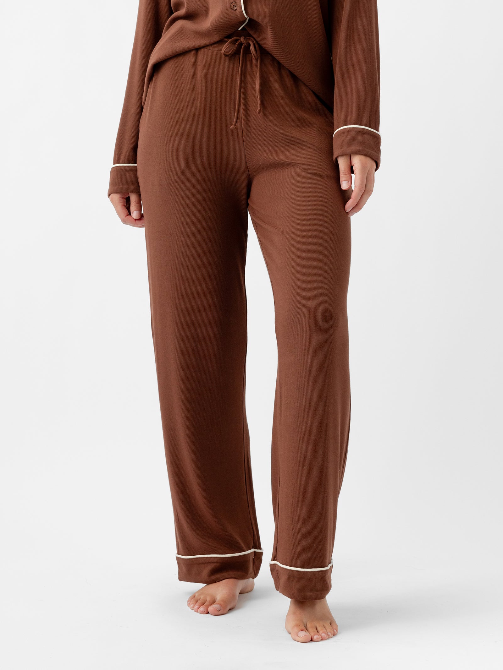 A person is standing barefoot on a light surface, wearing the Women's Bamboo Rib Knit Classic Pajama Pant from Cozy Earth in brown with white piping at the cuffs and a matching top. The outfit appears to be comfortable loungewear. The image is cropped to show the person from the waist down. 