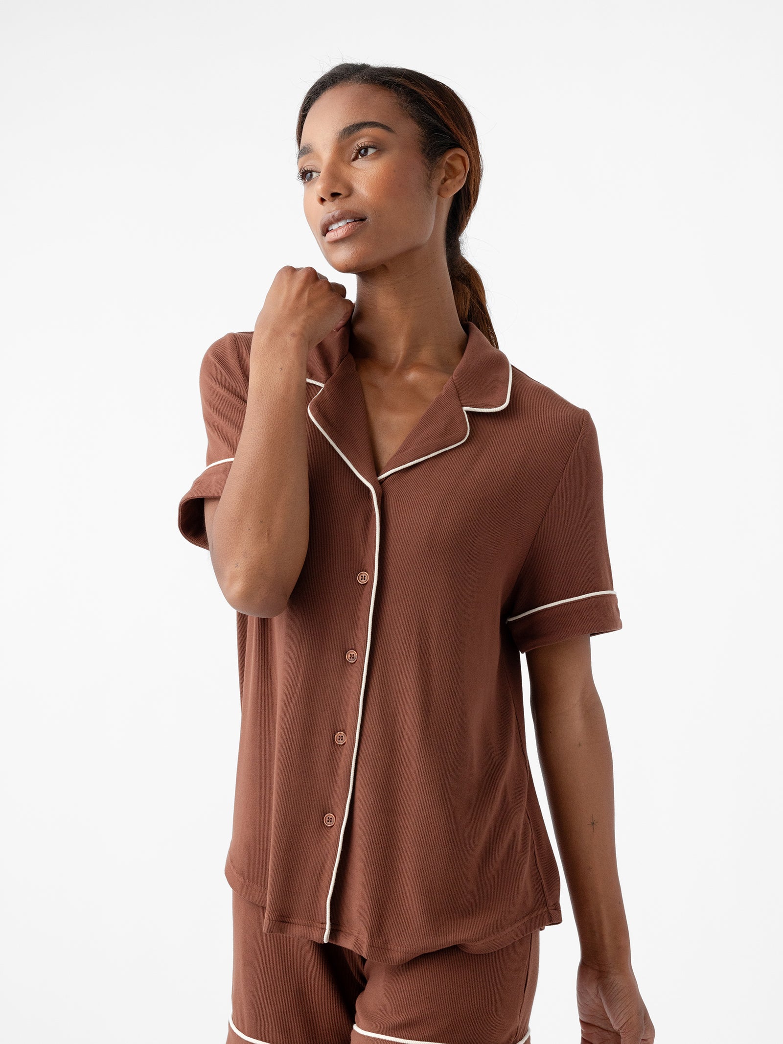 A person with long brown hair is standing against a white background. They are wearing the Cozy Earth Women's Bamboo Rib Knit Classic Short Sleeve Pajama Top in brown, featuring white piping along the edges. Their left hand is raised to touch their neck, and they have a relaxed, thoughtful expression on their face. 