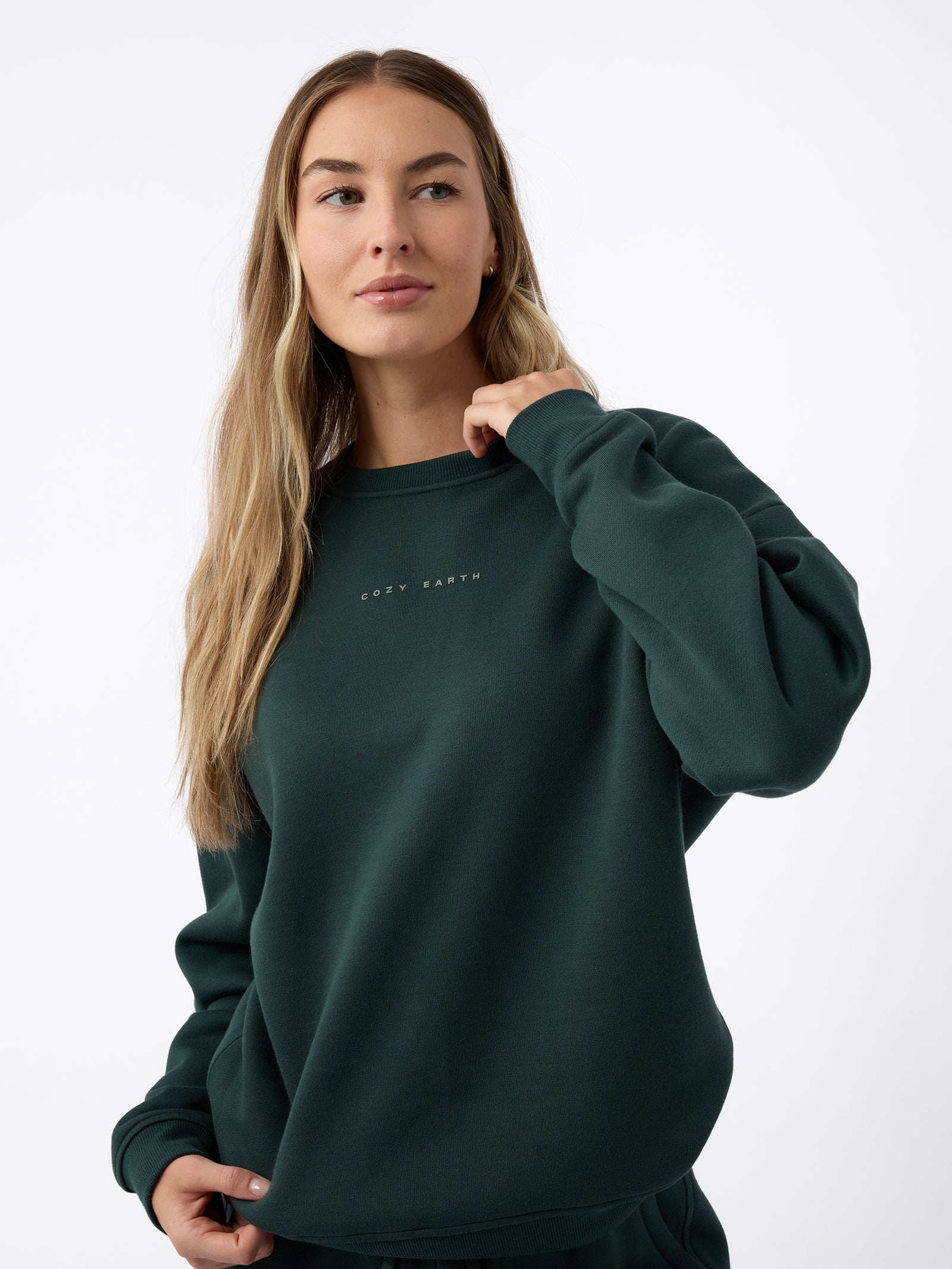 A person with long, light brown hair is wearing a dark green Women's CityScape Crewneck by Cozy Earth. They are looking slightly to the side, with one hand resting on the neckline of the sweatshirt. The background is plain white. 