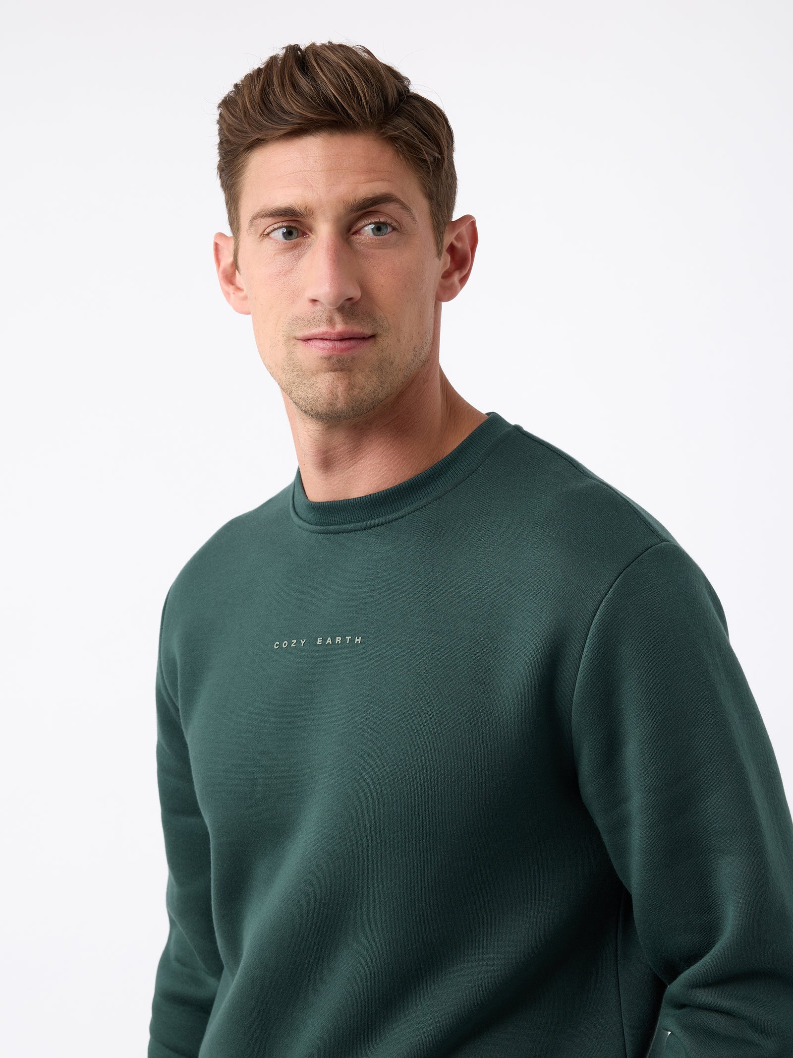 A man with short brown hair is wearing a dark green Men's CityScape Crewneck from Cozy Earth, featuring the words "COLD EARTH" printed on it. He is gazing slightly to the side against a plain white background. 