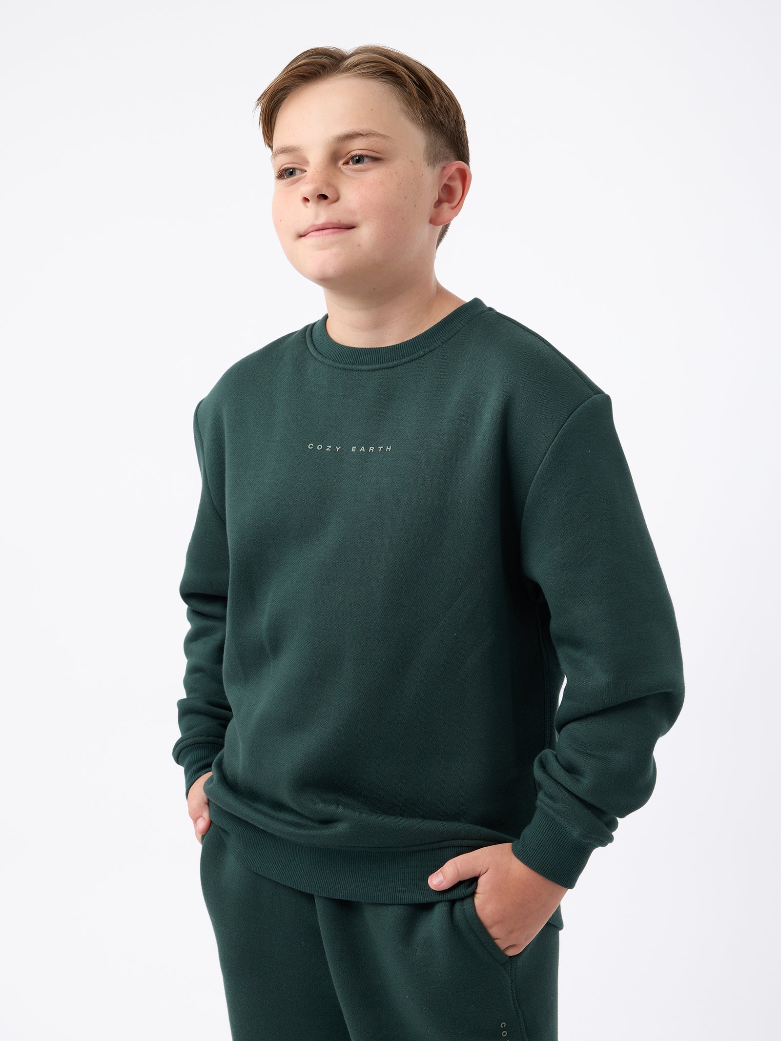 A boy wearing a Kid's CityScape Crewneck by Cozy Earth in dark green, along with matching pants, stands with hands in pockets against a plain white background. 