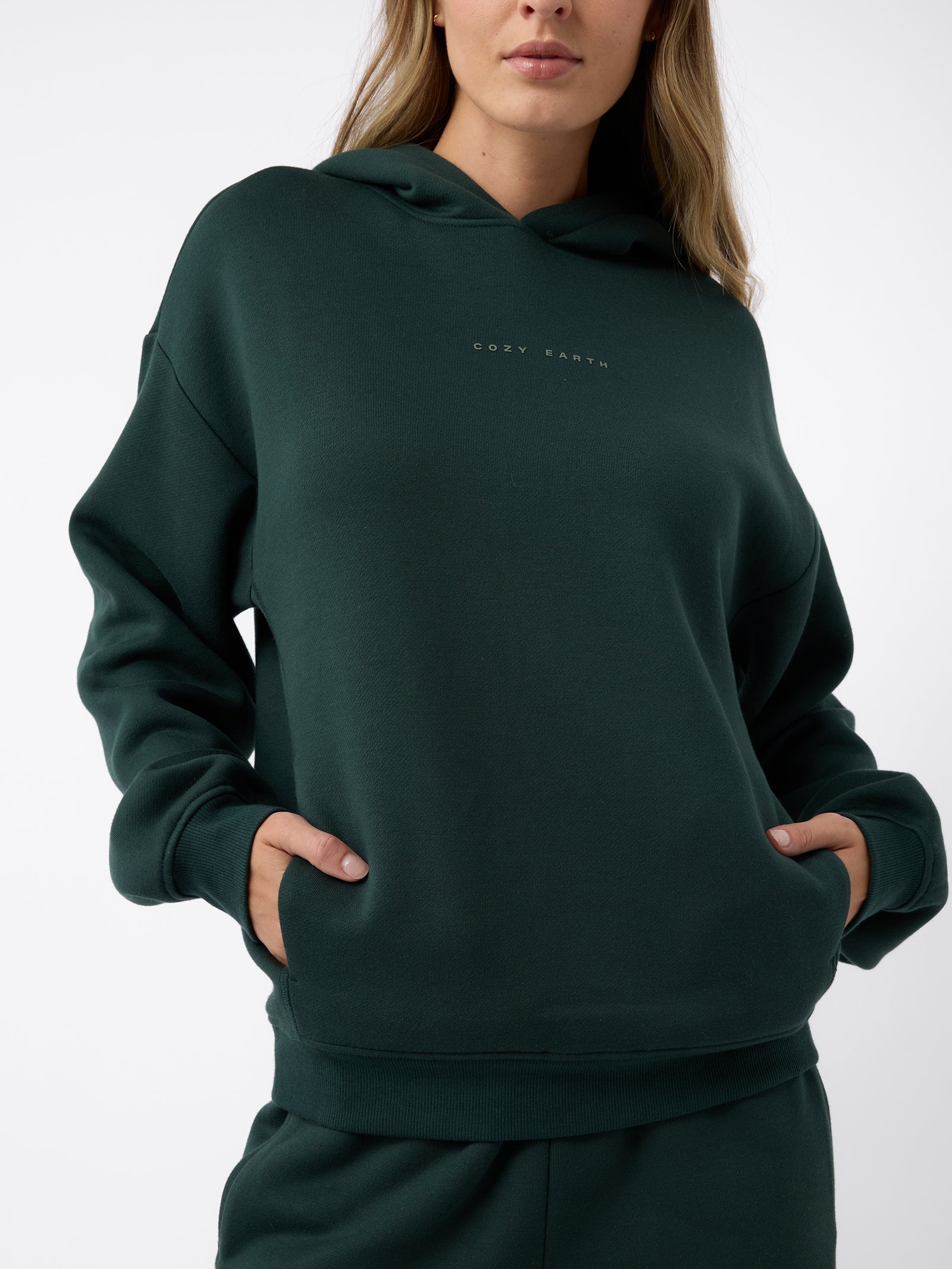 A woman wearing a Women's CityScape Hoodie by Cozy Earth, featuring the brand's name on the front, stands with her hands in her pockets against a white background. 