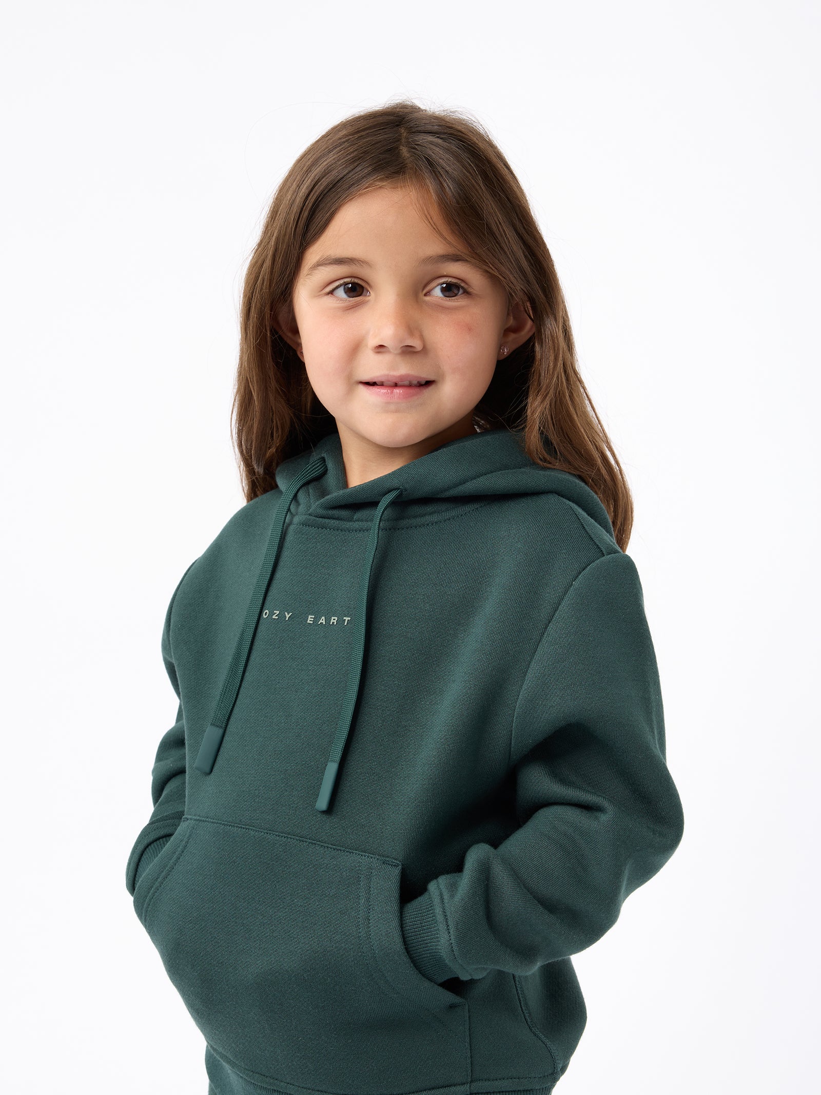 A young girl with long brown hair is wearing a Kid's CityScape Hoodie by Cozy Earth, featuring the brand logo. She has her hands in the hoodie pockets and is smiling slightly, standing against a plain white background. 