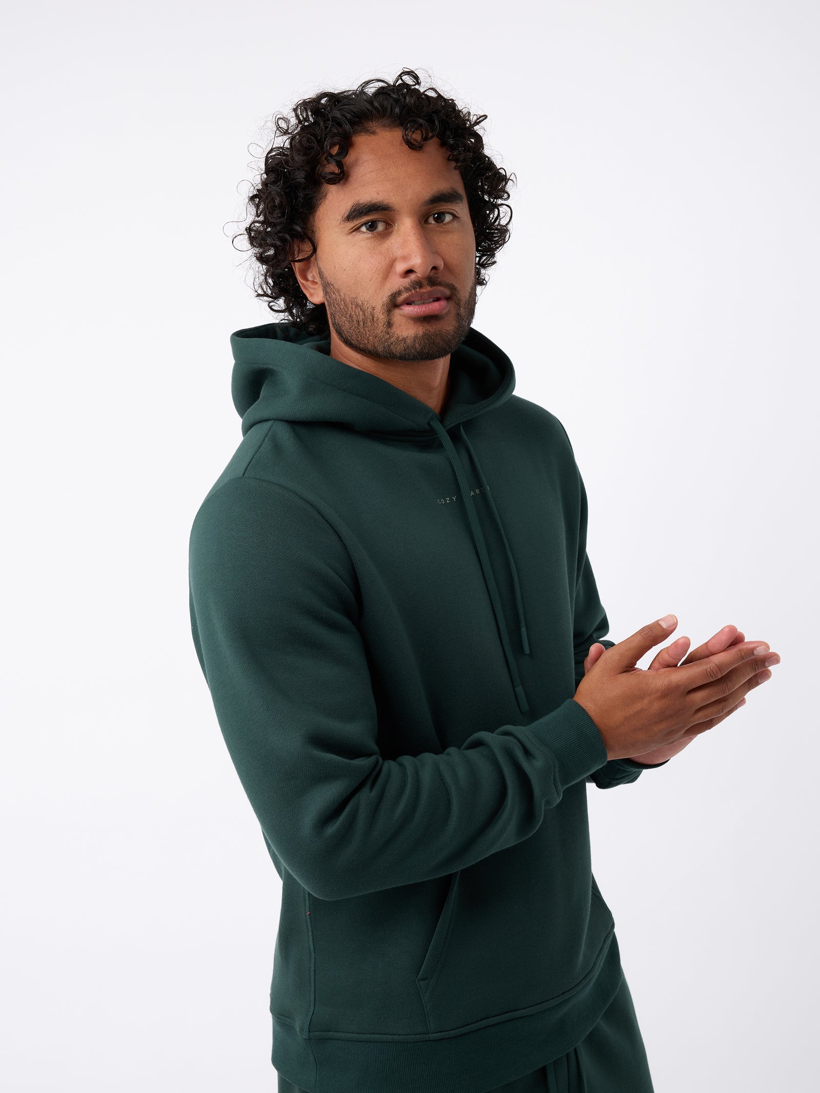 A person with curly hair is wearing a Cozy Earth Men's CityScape Hoodie in dark green and standing against a plain white background, with their hands together. 