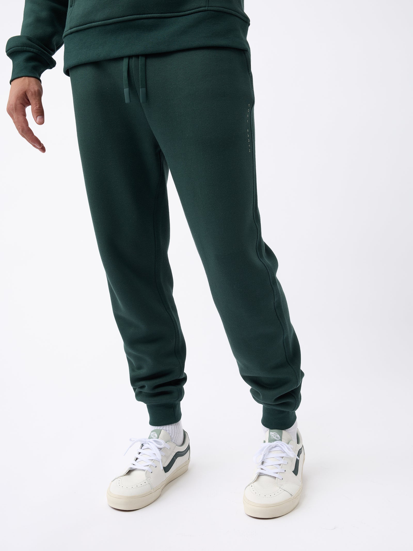 A person dressed in Cozy Earth's Men's CityScape Jogger in dark green and white sneakers is standing against a plain white background. The upper body is partially visible, showcasing a matching green sweatshirt. 