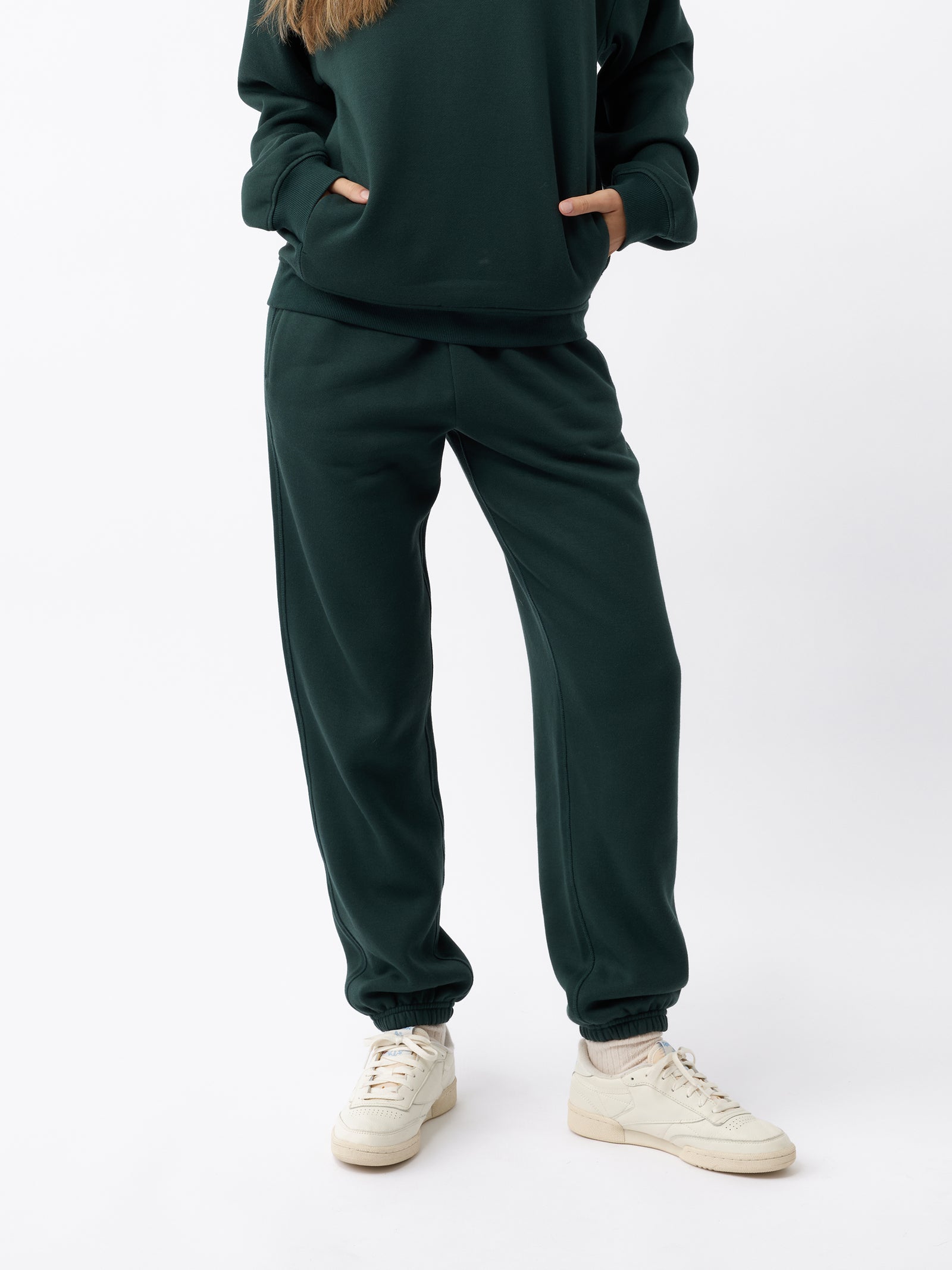 A person dressed in a dark green women's CityScape Sweatpant by Cozy Earth, with their hands in the pockets of this long-sleeve top and matching pants ensemble. They are wearing light-colored sneakers and standing against a plain white background. 