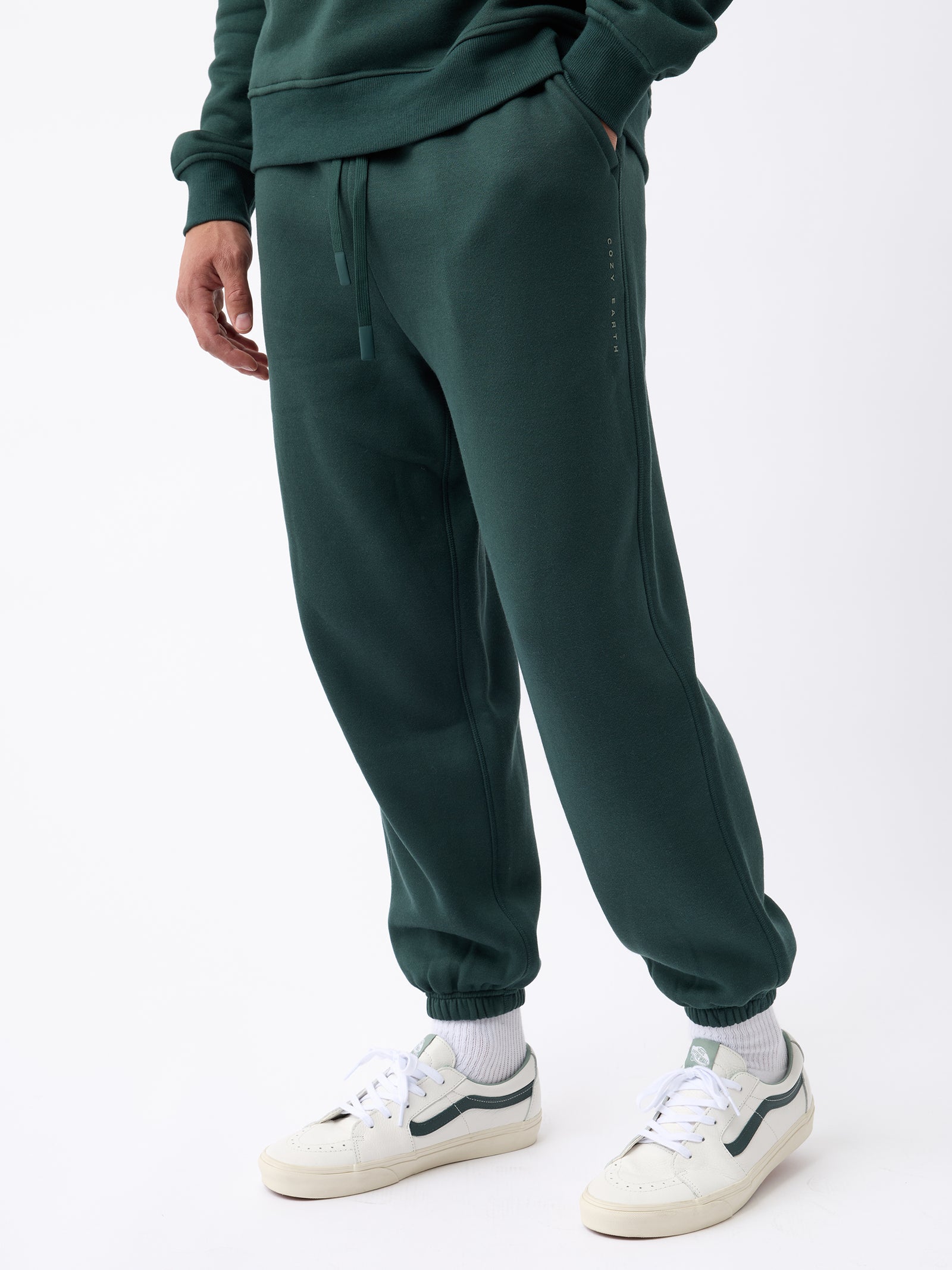 A person wearing Cozy Earth's Men's CityScape Sweatpant in dark green, paired with a matching sweatshirt. They are also sporting white sneakers featuring dark accents and white socks. The individual strikes a casual pose with their hands in pockets, set against a plain white background. 