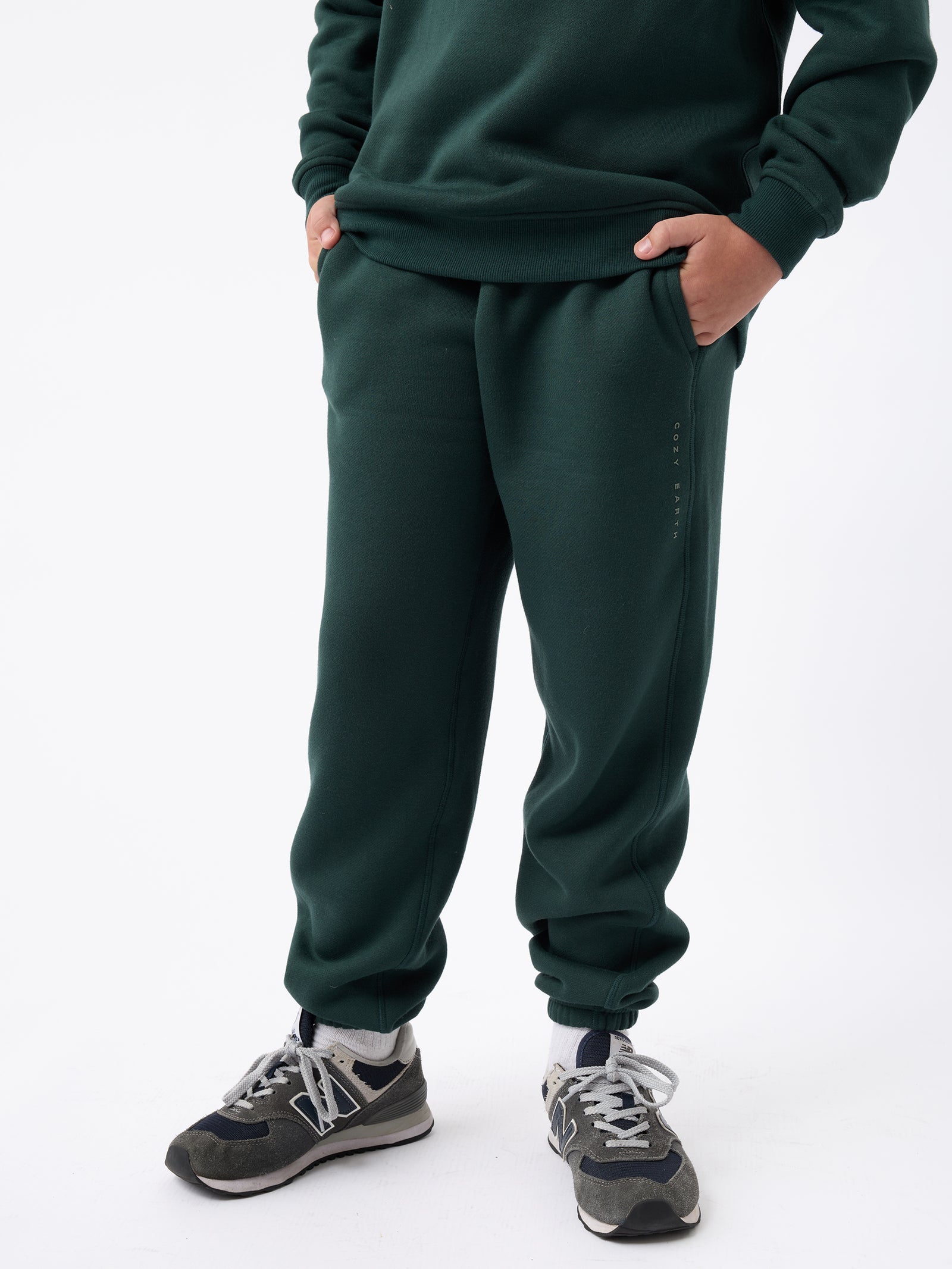 A person wearing Cozy Earth's Kid's CityScape Sweatpant in dark green stands against a plain white background with hands in pockets, paired with black and white sneakers. 