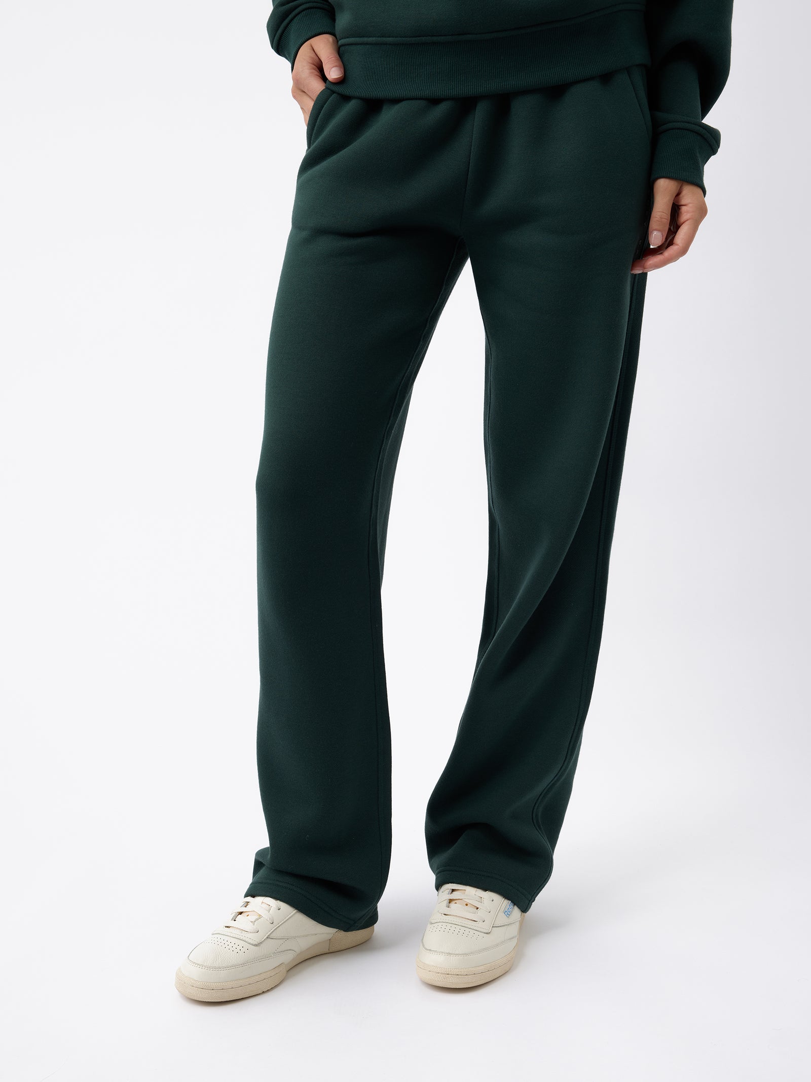 A person dressed in Cozy Earth's Women's CityScape Wide Leg Pant in a dark green shade, along with a matching hoodie, stands against a plain white background. White sneakers complement the outfit, and their hands are tucked into the pockets of the pants. 