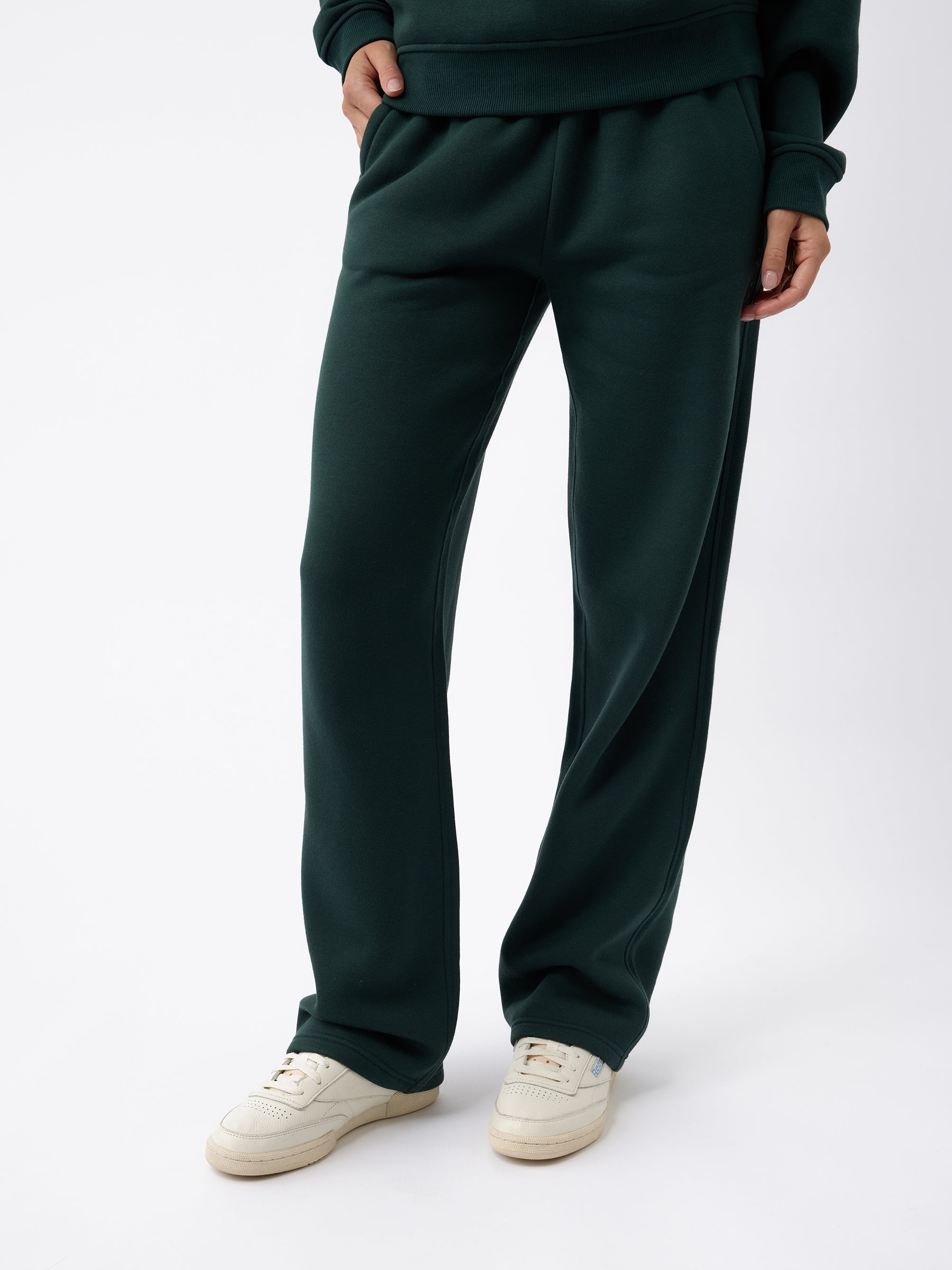 A person dressed in Cozy Earth's Women's CityScape Wide Leg Pant in a dark green shade, along with a matching hoodie, stands against a plain white background. White sneakers complement the outfit, and their hands are tucked into the pockets of the pants. |Color:Spruce