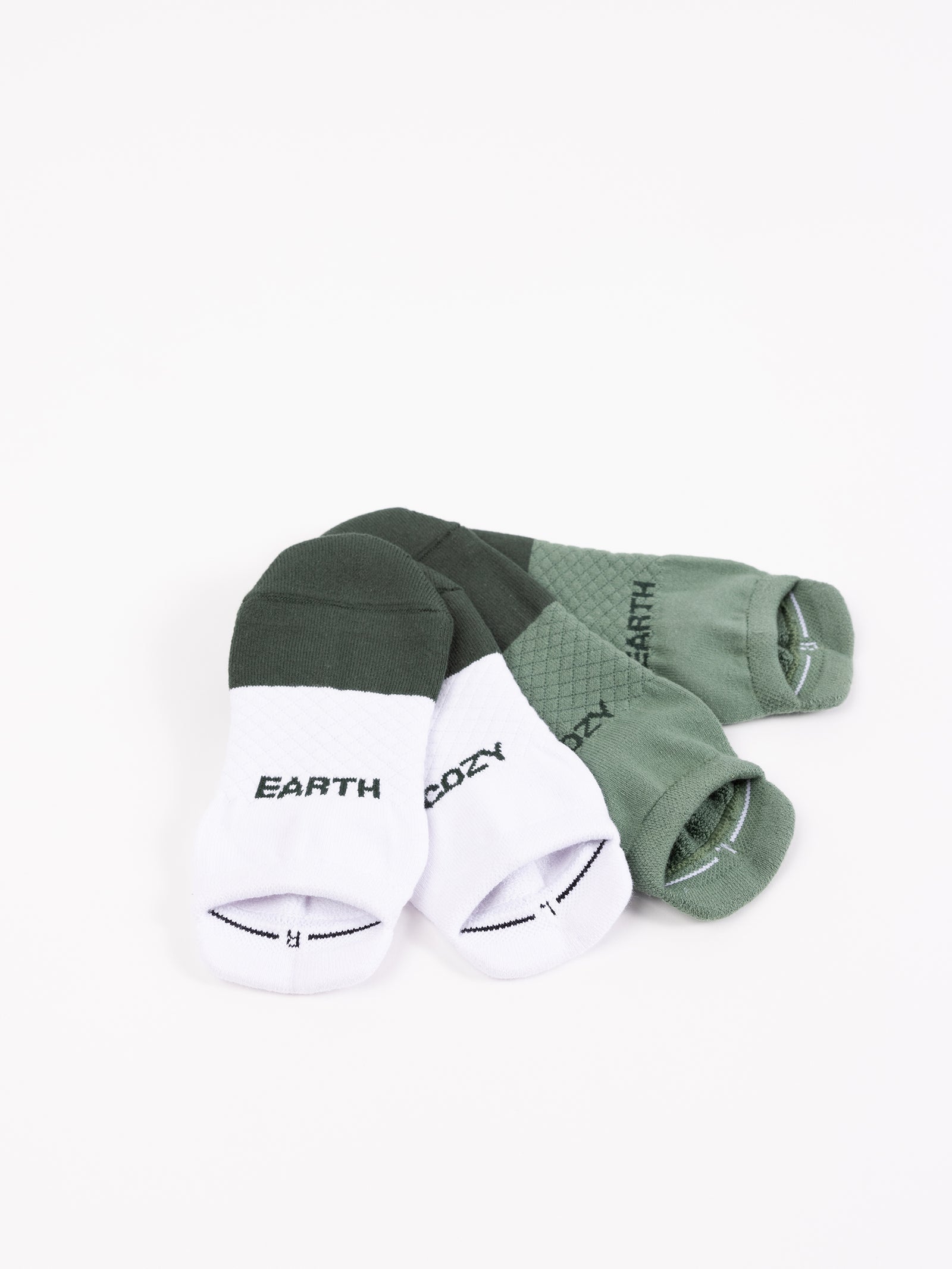 Three pairs of socks are arranged on a white background. Two pairs are from the "Essential Ankle Sock 2-Pack" by Cozy Earth, featuring white bottoms with "EARTH" and "COZY" written on them in black and green tops. The third pair is entirely green with alternating textured and plain patterns. 