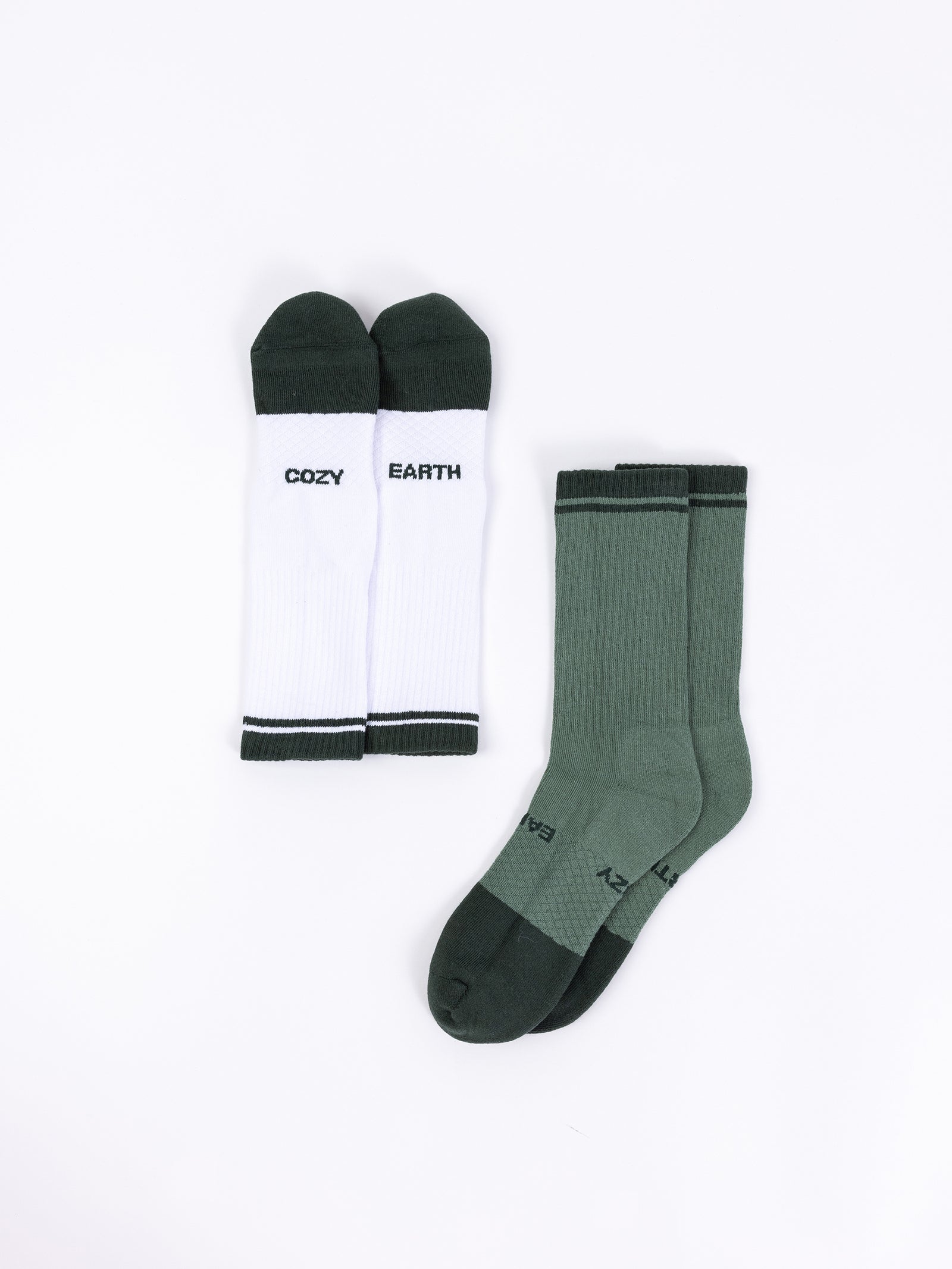 Two pairs of socks from the Essential Calf Sock 2-Pack by Cozy Earth are pictured on a white background. The left pair is white with black toes and heels, featuring "COZY" and "EARTH" written in black on each sock. The right pair is green with darker green toes, heels, and cuffs. 