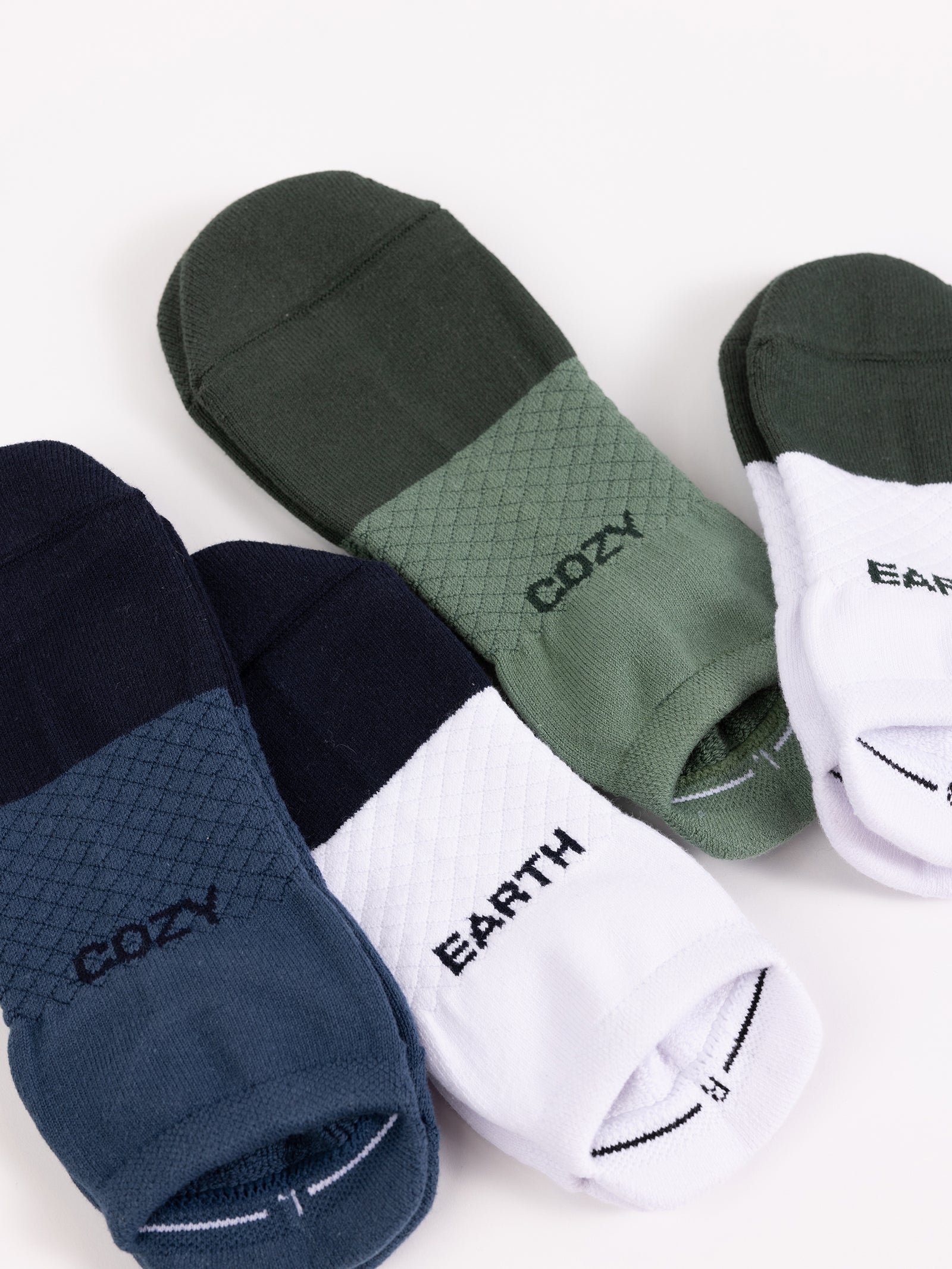 Two pairs of Essential Ankle Socks from Cozy Earth lie on a white surface. One pair boasts a black and gray design with "COZY" written on them, while the other features a green and white design with "EARTH" written at the top. 