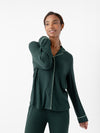 A person wearing Cozy Earth's Women's Bamboo Rib Knit Classic Long Sleeve Pajama Top in dark green with light piping details stands against a plain white background. They are smiling and have one hand relaxed on their neck. The pajama top features long sleeves and buttons down the front. |Color:Spruce