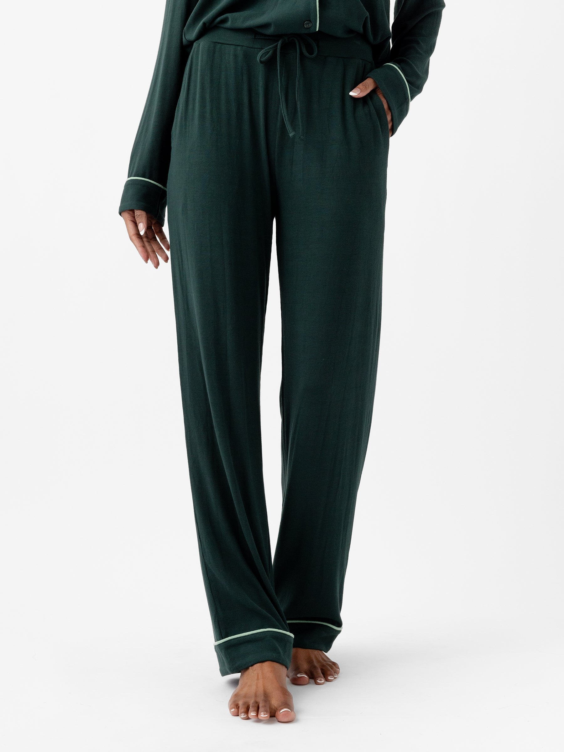 A person is wearing Cozy Earth's Women's Bamboo Rib Knit Classic Pajama Pant in dark green, featuring white trim around the cuffs. The loose-fitting pants are tied at the waist with a drawstring, and the person's hands rest in their pockets. They are standing barefoot, with the image focused from the shoulders down. |Color:Spruce