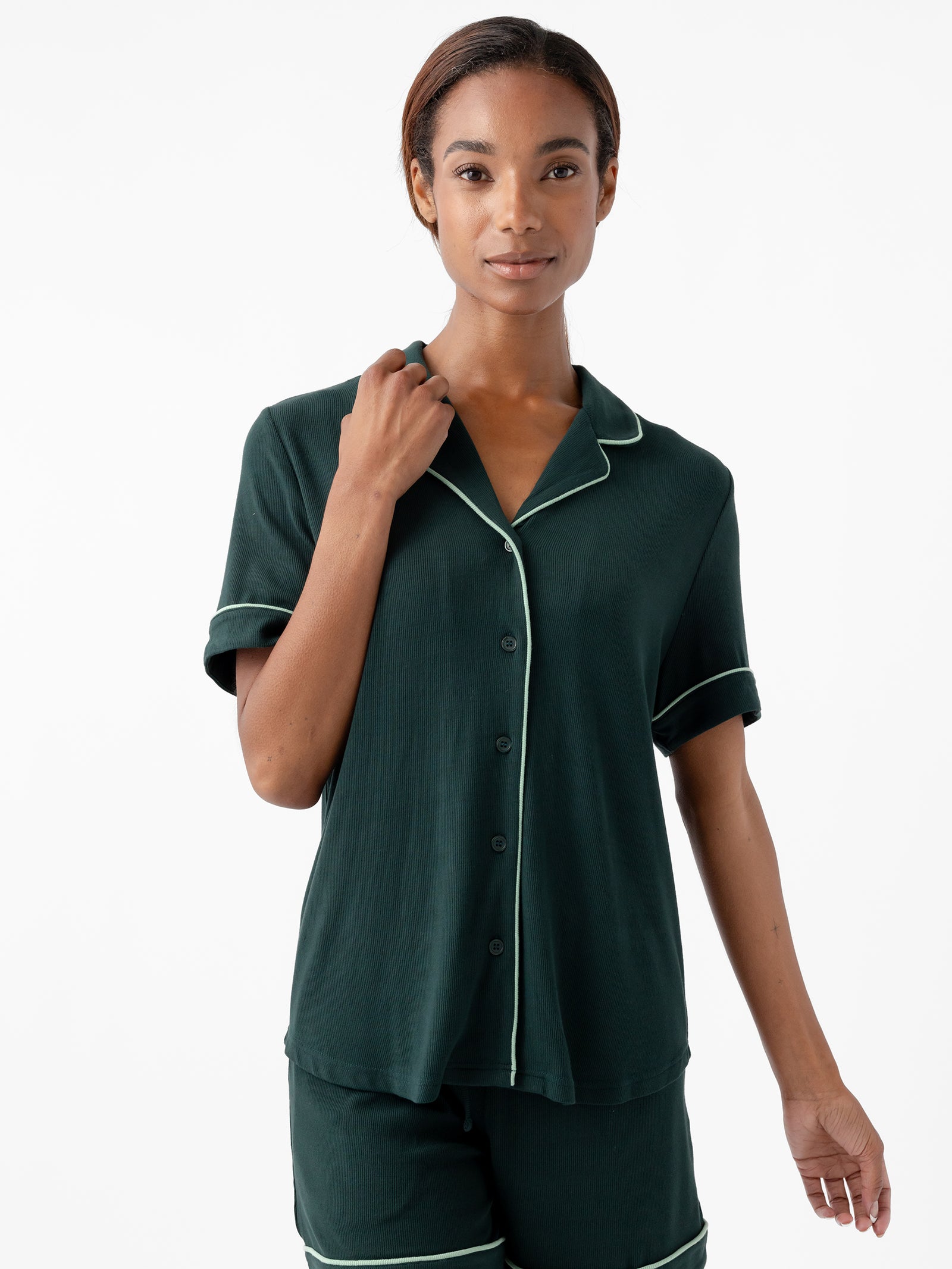 A person with dark hair is wearing a dark green Women's Bamboo Rib Knit Classic Short Sleeve Pajama Top by Cozy Earth, featuring a button-up design with a collared neckline and short sleeves, accented with light-colored piping. They are standing against a plain white background and looking towards the camera. 