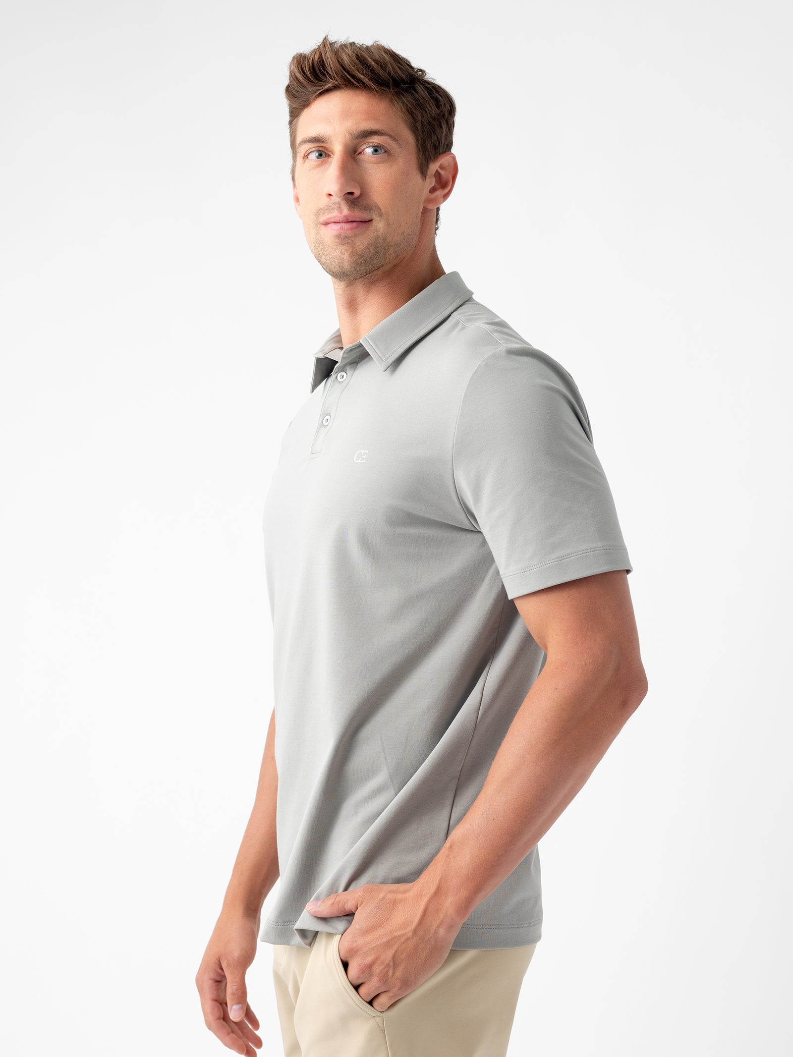 A person with short, light brown hair is wearing the Men's Everyday Polo by Cozy Earth in a light gray shade, paired with light beige pants. They are standing and slightly looking over their left shoulder with a neutral expression against a plain white background. 