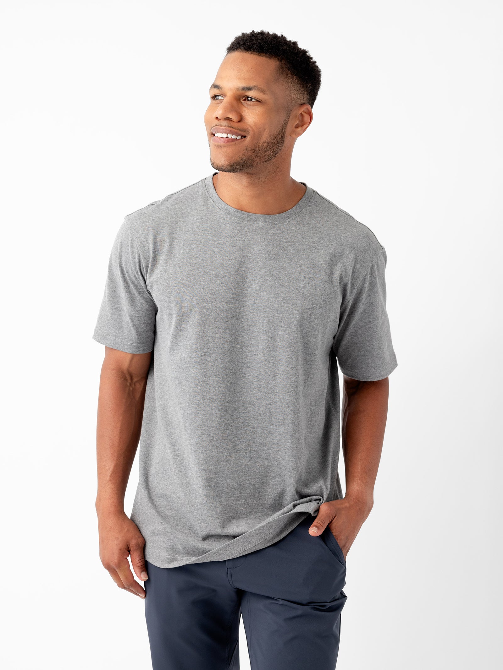 A man stands against a white background, smiling and looking to the side. He is dressed casually in a plain gray Men's All Day Tee by Cozy Earth and dark blue pants. One hand is in his pocket, while the other hangs by his side. 