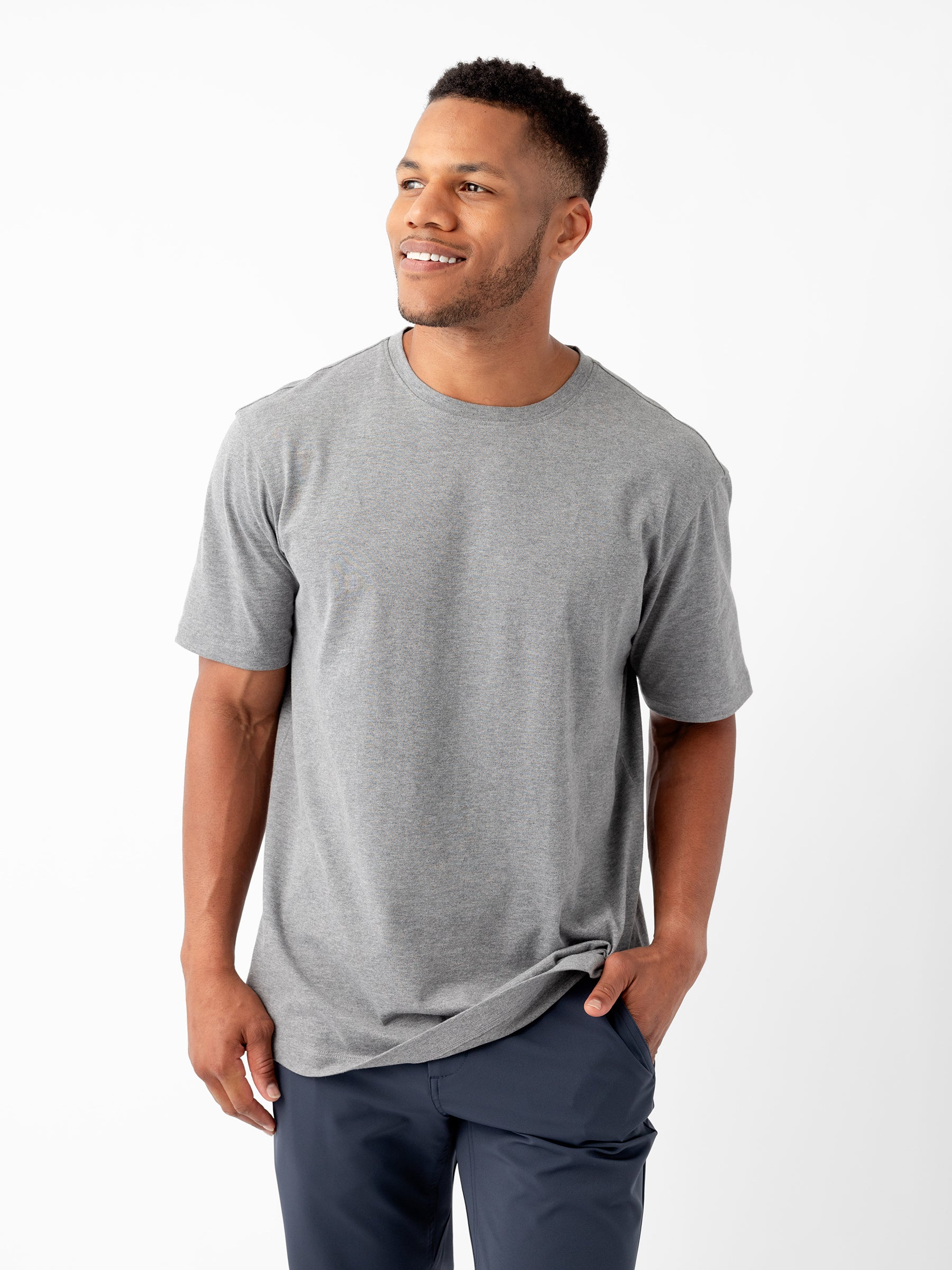 A man stands against a white background, smiling and looking to the side. He is dressed casually in a plain gray Men's All Day Tee by Cozy Earth and dark blue pants. One hand is in his pocket, while the other hangs by his side. |Color:Stone Heather