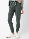 A person is standing against a plain background, wearing Cozy Earth's Women's Studio Jogger in dark green with a drawstring waist, paired with a light green sweatshirt. They are also wearing white sneakers. Their right hand is resting on their hip. |Color:Storm