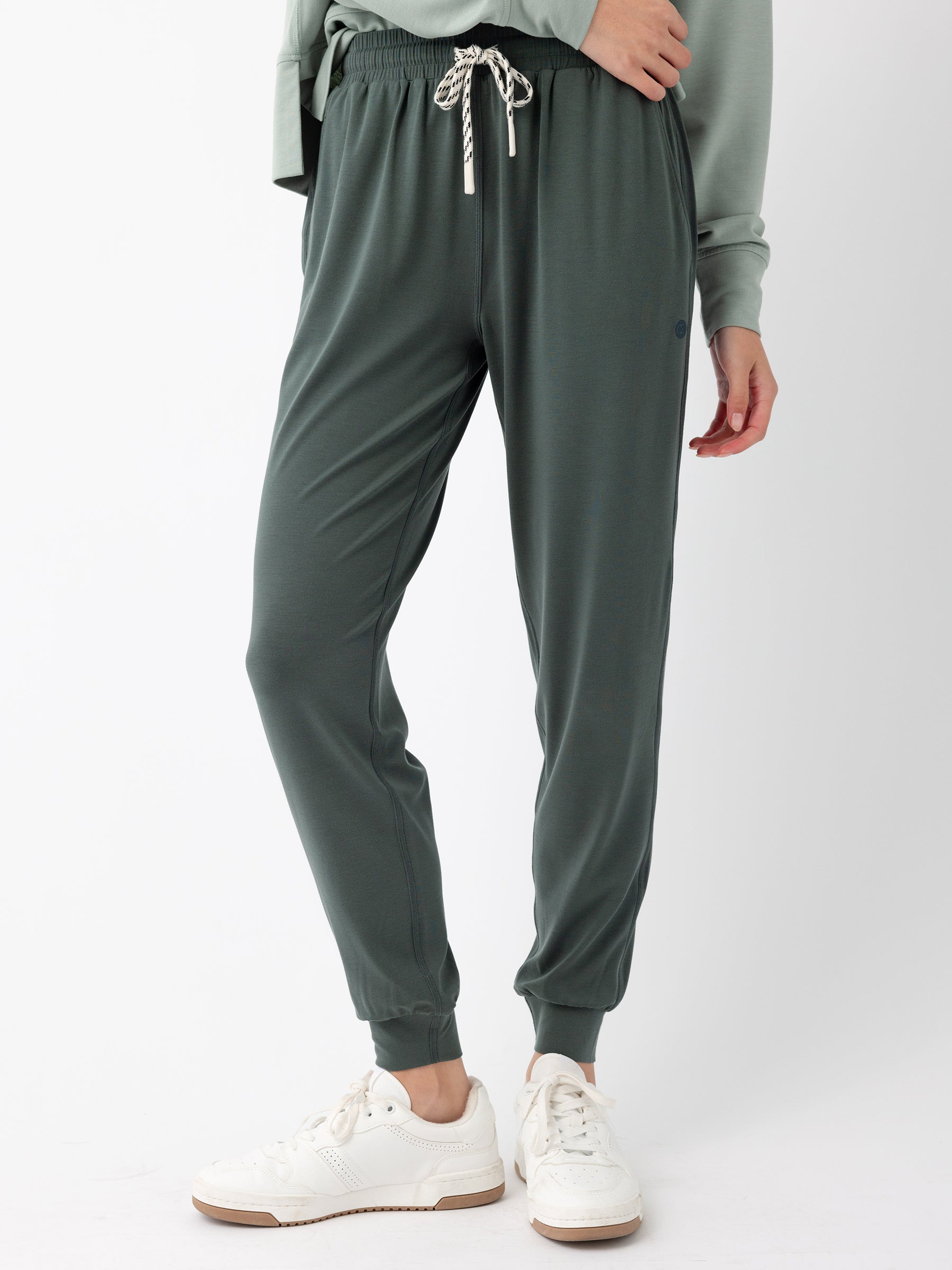 A person is standing against a plain background, wearing Cozy Earth's Women's Studio Jogger in dark green with a drawstring waist, paired with a light green sweatshirt. They are also wearing white sneakers. Their right hand is resting on their hip. |Color:Storm