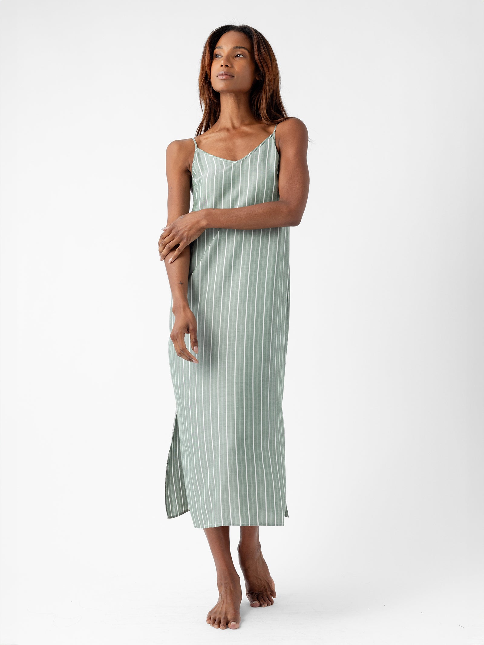 A person stands barefoot wearing the Women's Soft Woven Nightgown by Cozy Earth, featuring a sleeveless design with green stripes and side slits. The background is plain white. The person's arms are relaxed, and they appear thoughtful. 