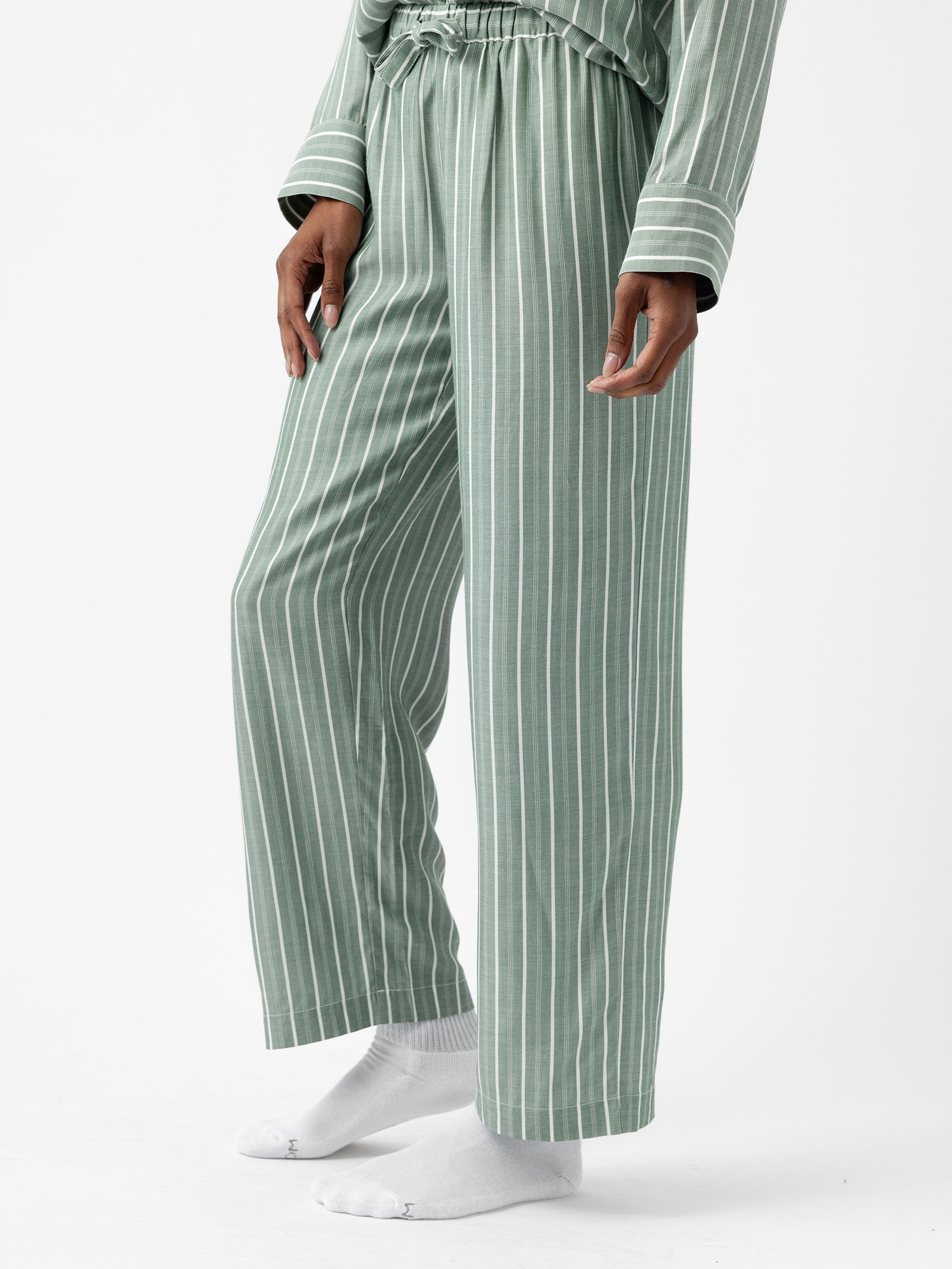 A person wearing the Women's Soft Woven Pajama Pant by Cozy Earth, featuring a green and white striped pattern, is standing on a white surface. The pajama pants have a loose fit and are paired with white socks, while the person's hands are relaxed by their sides. 