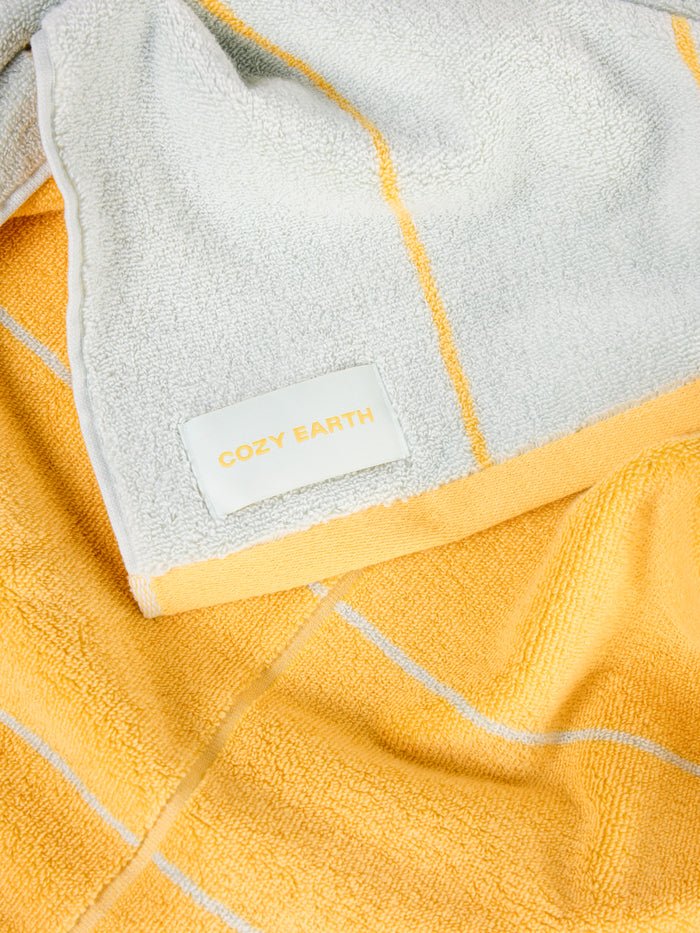 A close-up of the Windowpane Resort Towel in yellow and gray, featuring a checkered pattern. The inner label shows "Cozy Earth" in gold letters. The fabric looks soft and plush. 