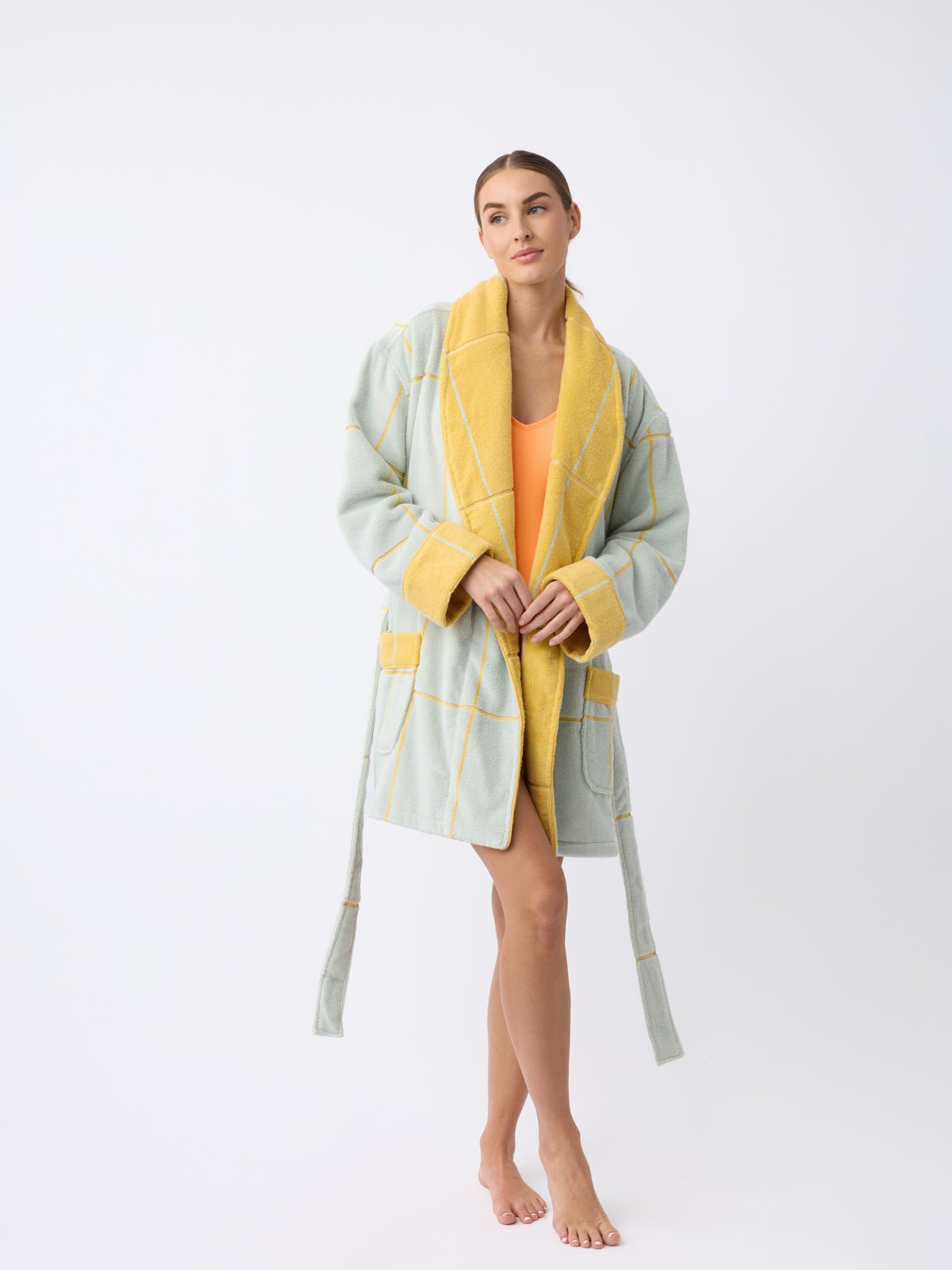 A woman models Cozy Earth's Windowpane Resort Robe, featuring a yellow and gray checkered pattern, over an orange garment. They stand barefoot on a plain white background, holding the robe's lapel with one hand. |Color:Sunny Sky