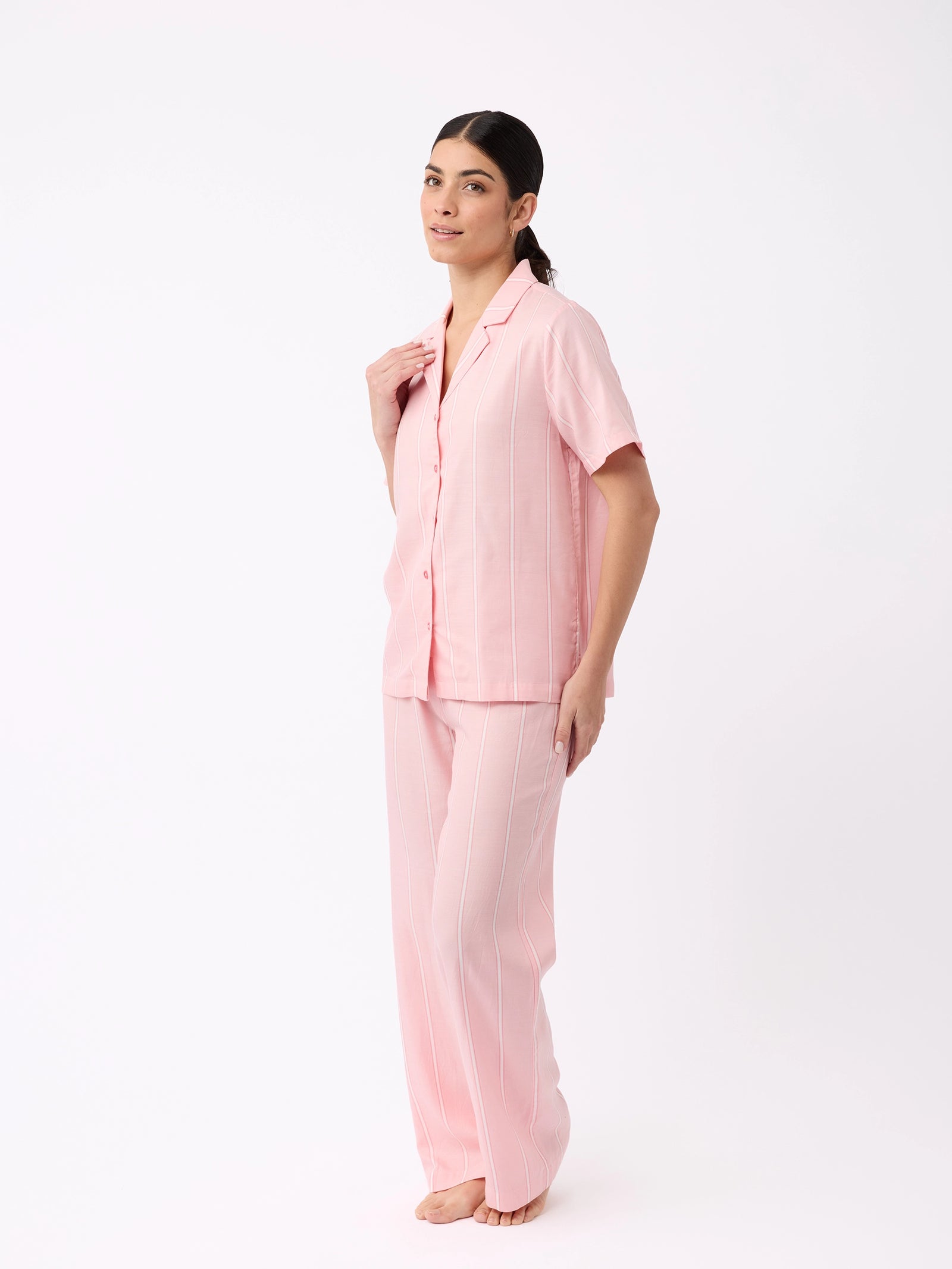 A person with long dark hair tied back smiles gently while standing barefoot against a white background, wearing Cozy Earth's Women's Soft Woven Pajama Pant set in pink with white pinstripes, featuring a matching short-sleeve button-up shirt and long pants. 