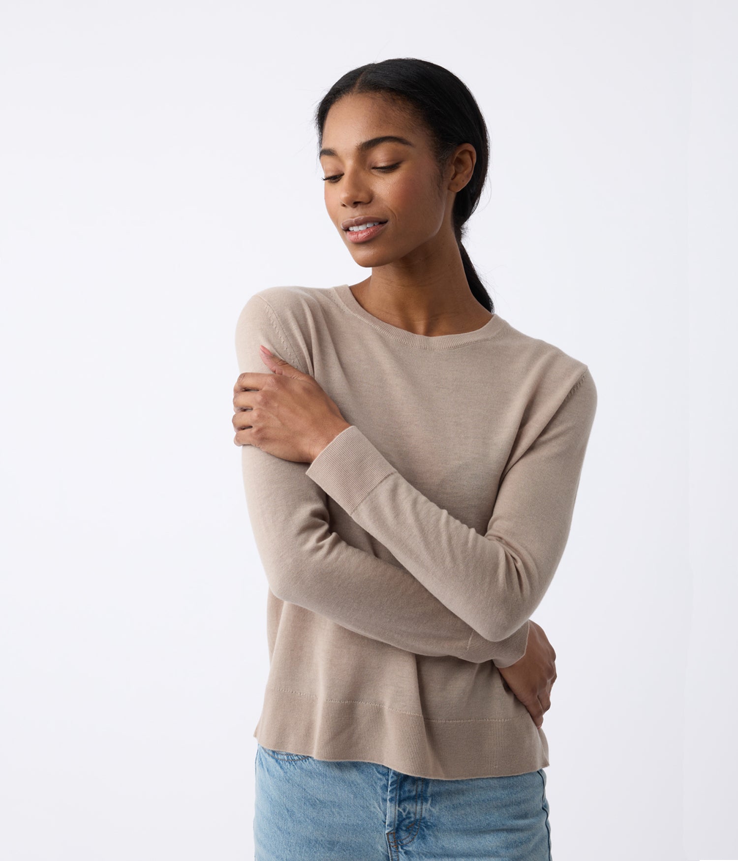 A woman in Cozy Earth's Women's AirKnit Crewneck Sweater, beige with long sleeves, stands against a plain backdrop. She has long dark hair, gently touches her arm with a serene expression, and gazes slightly downward while wearing light blue jeans. |Color:Taupe