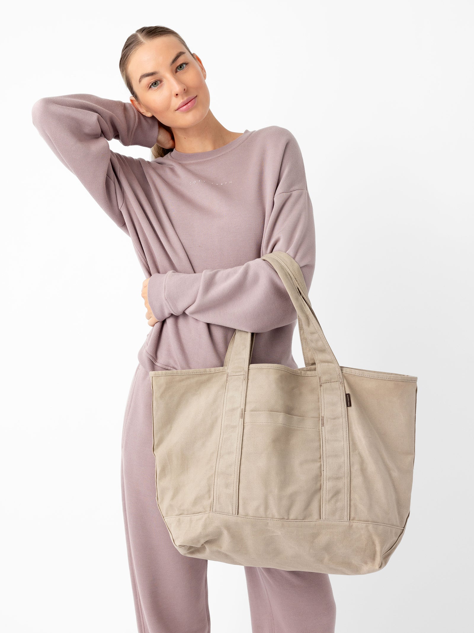 A person dressed in a lavender sweatsuit poses against a white background, resting their left arm on their head. They are holding a large Waxed Canvas Tote from Cozy Earth with both hands, displaying it toward the camera. 