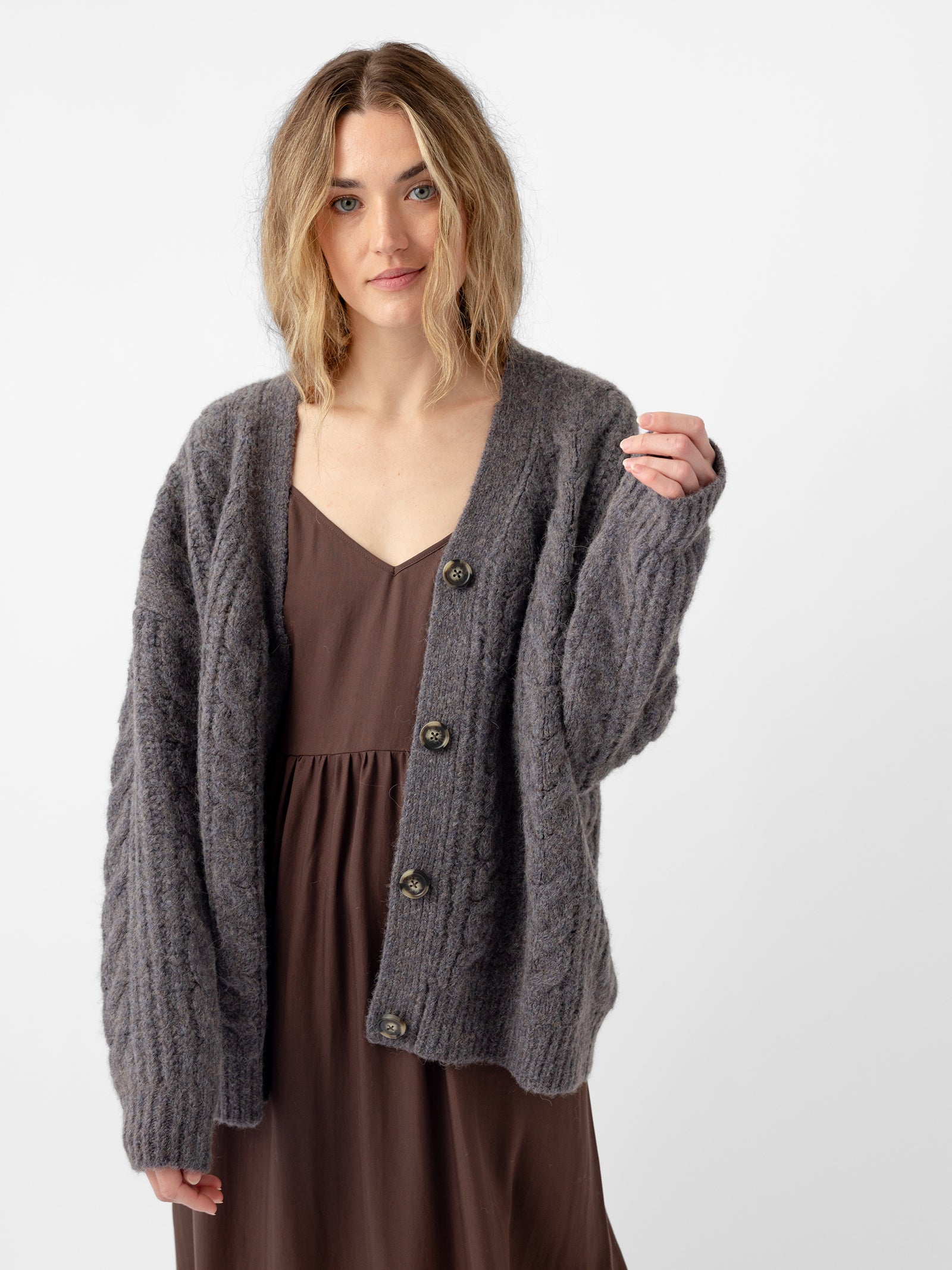 A person with wavy hair is wearing a dark brown dress and a Cozy Earth Oversized Cable Knit Cardigan in gray, featuring buttons. They stand against a plain white background, with a relaxed expression and slightly raised hand. 