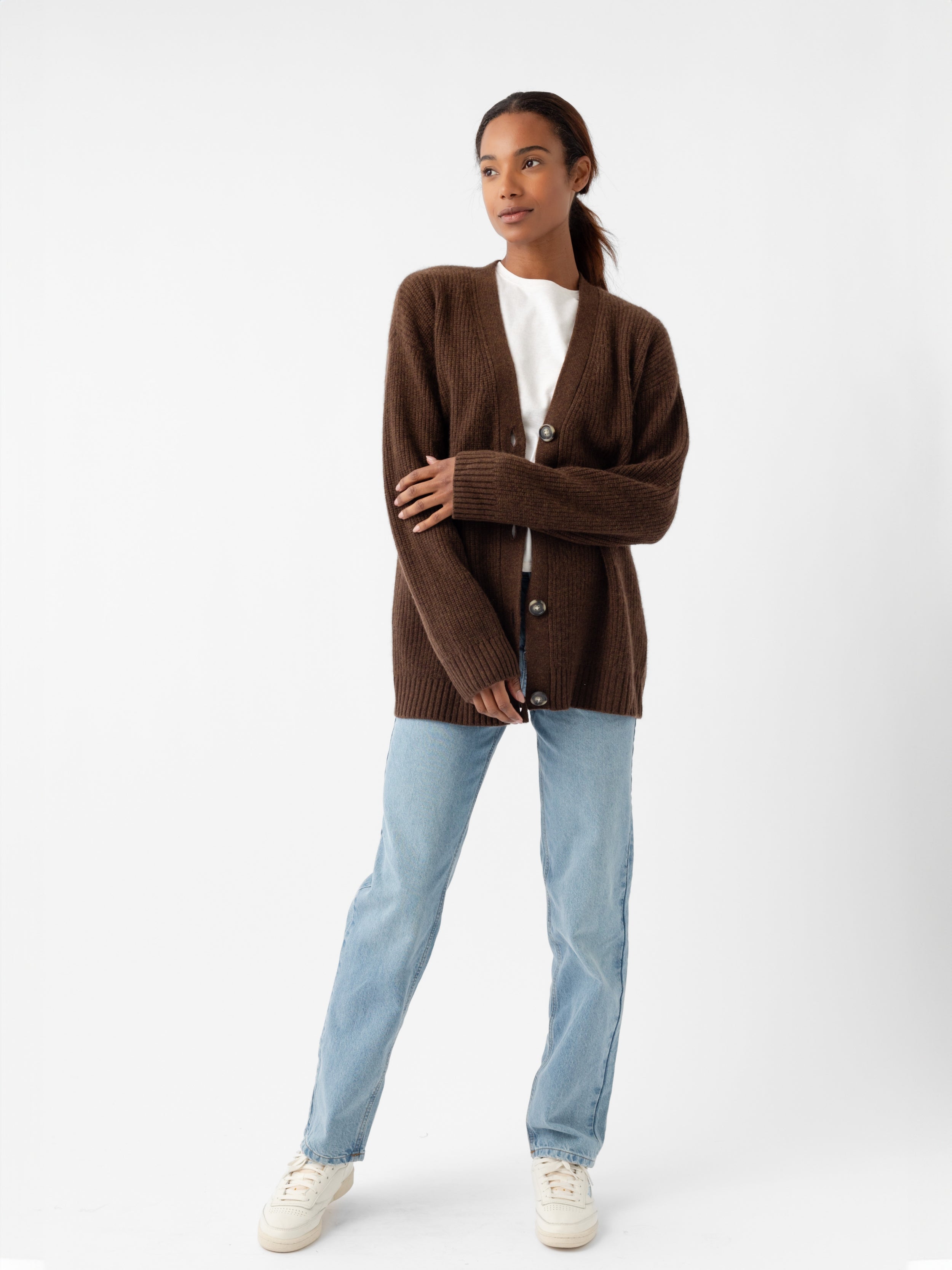 A person stands wearing an Oversized Sunday Cashmere Cardigan from Cozy Earth over a white shirt, paired with light blue jeans and white sneakers. They have long hair pulled back and are posed against a plain white background. |Color:Tobacco