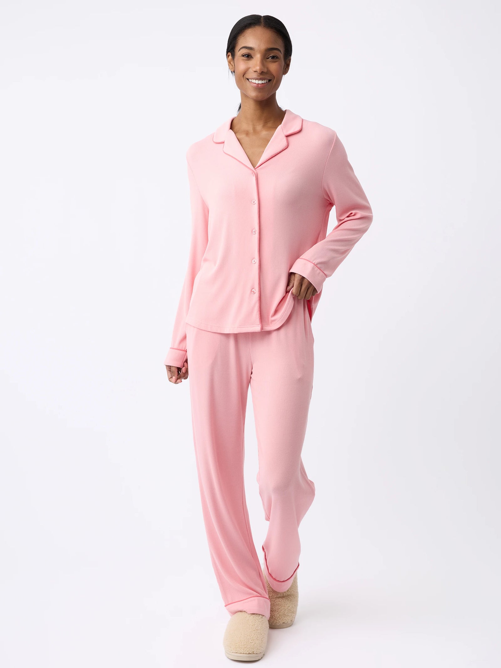 A woman is wearing Cozy Earth's Women's Bamboo Rib-Knit Classic Pajama Pant in light pink with a button-up top and beige slippers, smiling against a plain white background. 