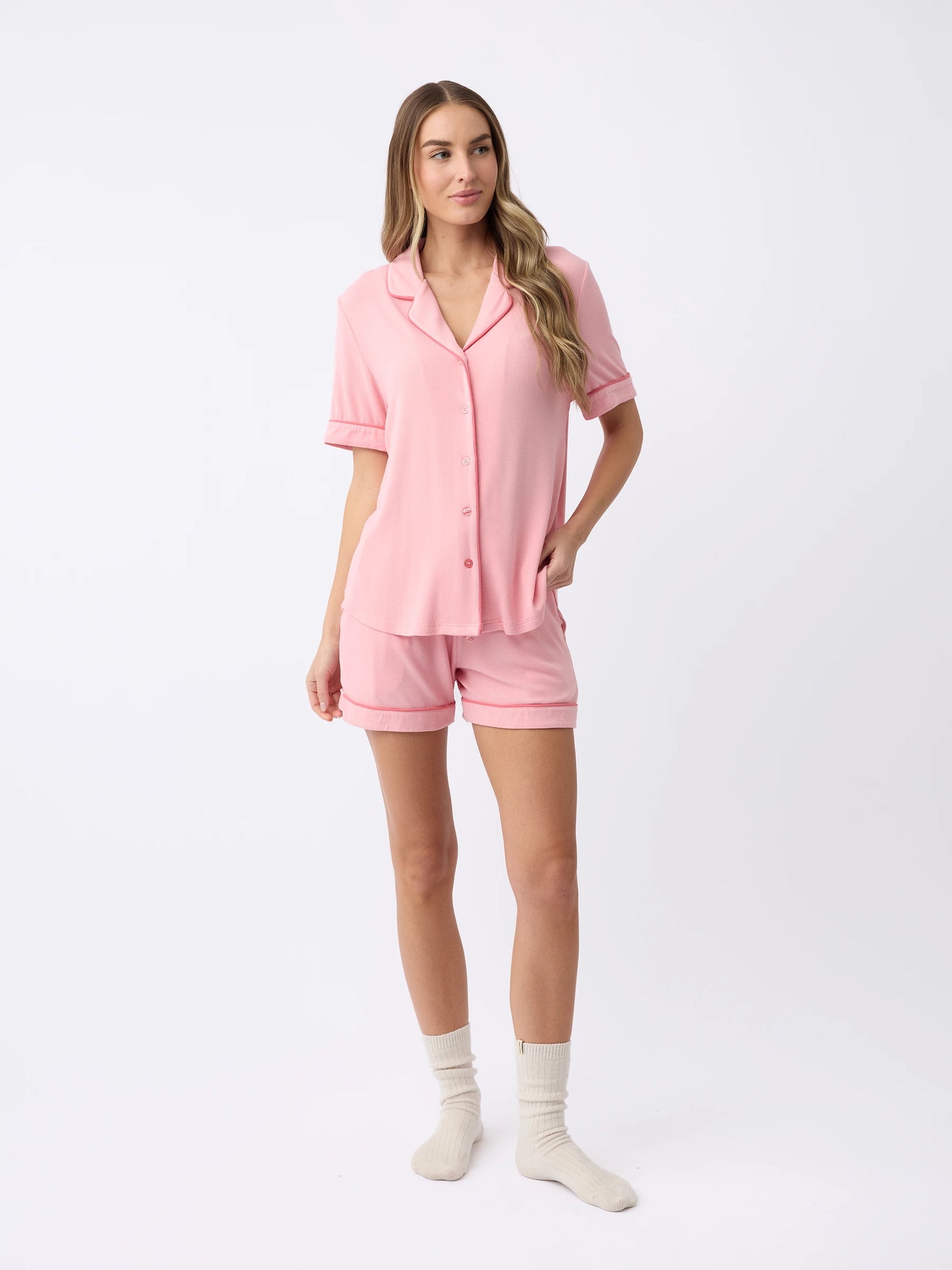 Against a plain white background, a woman models the Women's Rib-Knit Classic Short Sleeve Pajama Set by Cozy Earth. She wears the pink button-up top and matching shorts, paired with cream-colored socks. Her right hand rests in her pocket, and her long wavy hair cascades down. 