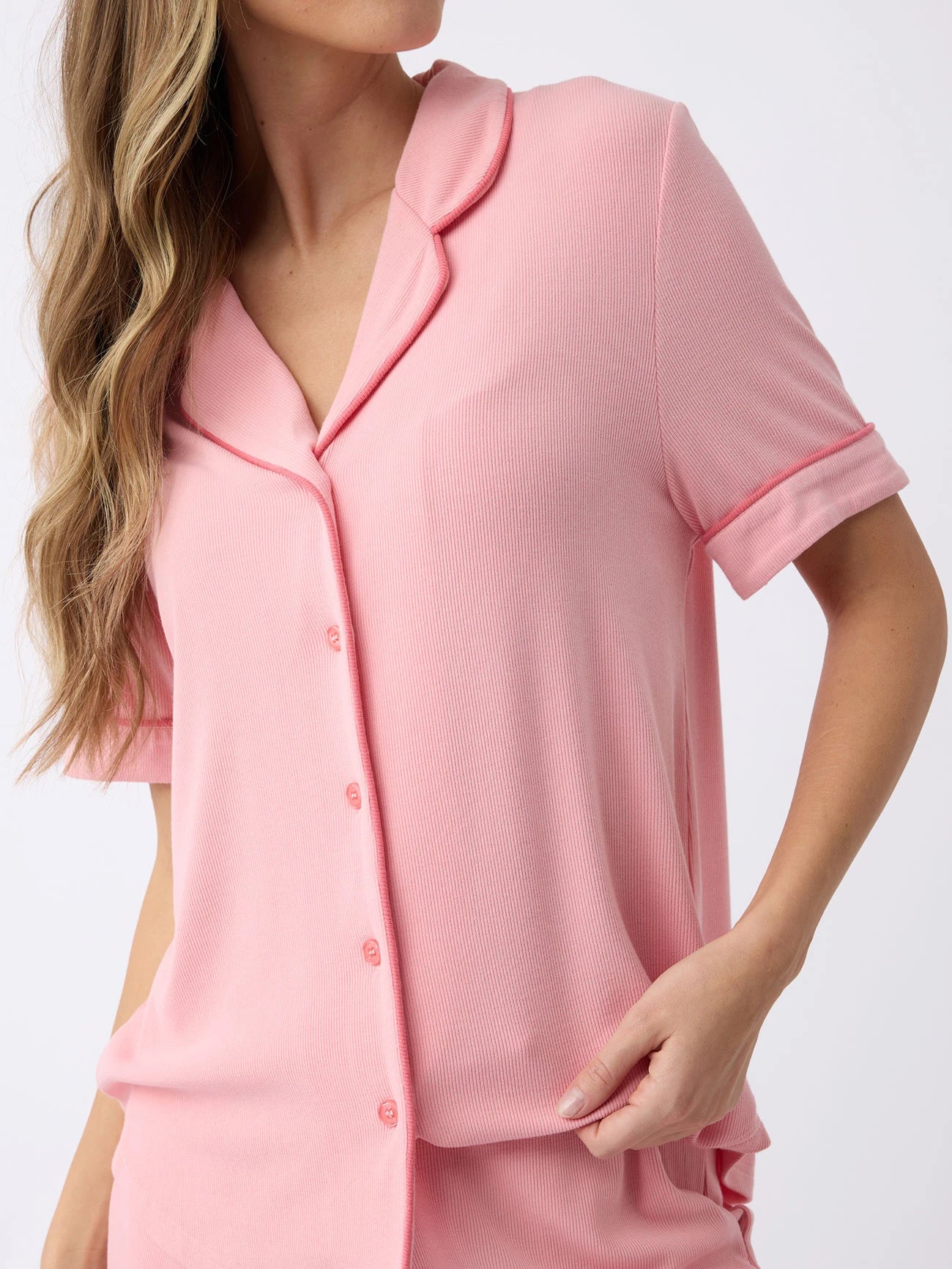 A woman with long, wavy hair in a plain white background wears a Cozy Earth Women's Bamboo Rib-Knit Classic Short Sleeve Pajama Top in light pink, featuring a button-up front and collar. She's gently touching the side of the top with one hand. |Color:Tulip