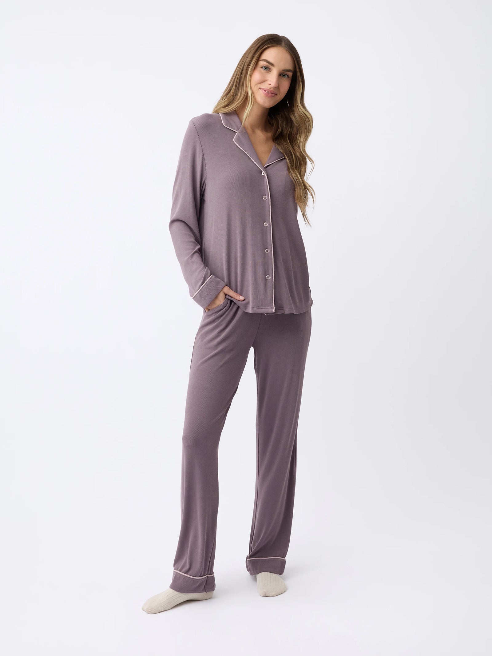 A woman stands in the Cozy Earth Rib-Knit Classic Long Sleeve Pajama Set, featuring a soft lavender tone with white piping, a button-up top, and long pants. They're barefoot against a plain white backdrop. 