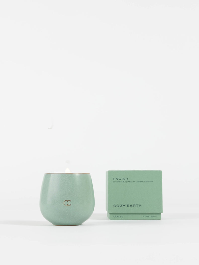 A pale green Cozy Earth Candle UNWIND in a round container, with a small flame, sits beside its matching box labeled "UNWIND" and "Cozy Earth" on a plain white background.