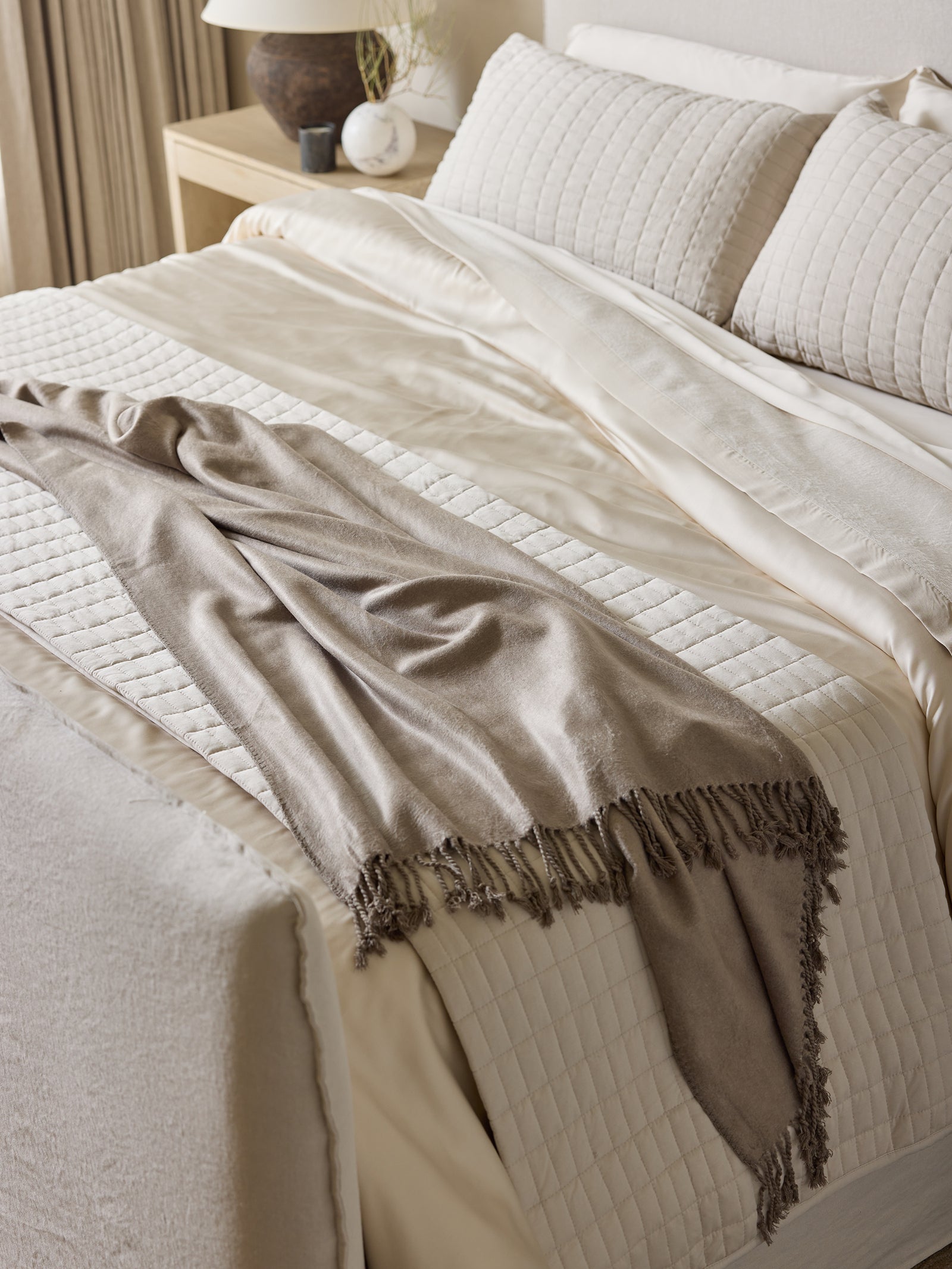 A neatly made bed with cream-colored sheets, a quilted blanket, and two pillows. The Bamboo Tassel Throw from Cozy Earth, featuring fringed edges, is casually draped across the lower part of the bed. The bedside table holds a lamp, a small plant, and some decorative items. 