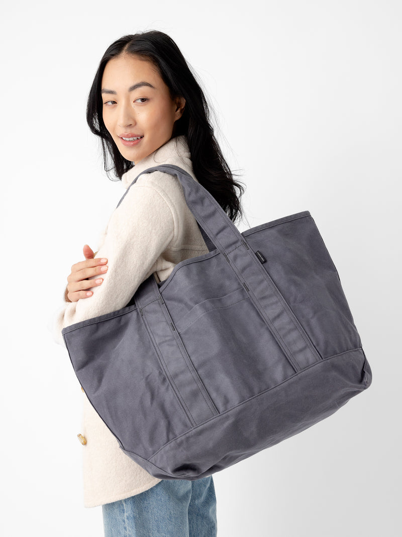 A person with long dark hair is wearing a light-colored coat and blue jeans. They have a large Cozy Earth Waxed Canvas Tote over their shoulder. The background is plain white.