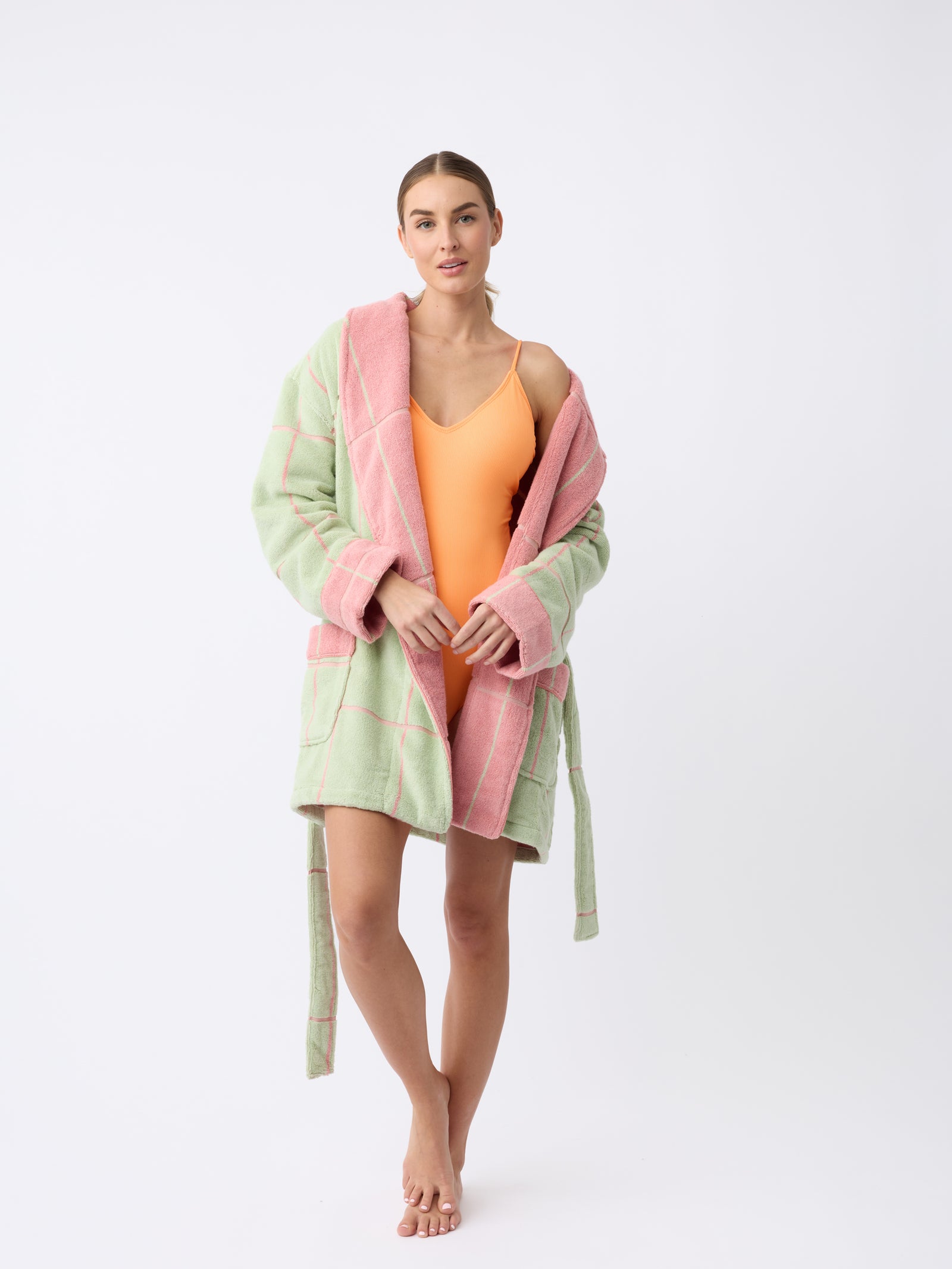 A person stands against a plain white background wearing the Windowpane Resort Robe by Cozy Earth, a pastel green and pink checkered robe. They're barefoot in an orange swimsuit, with a relaxed expression and holding the robe's edge. 