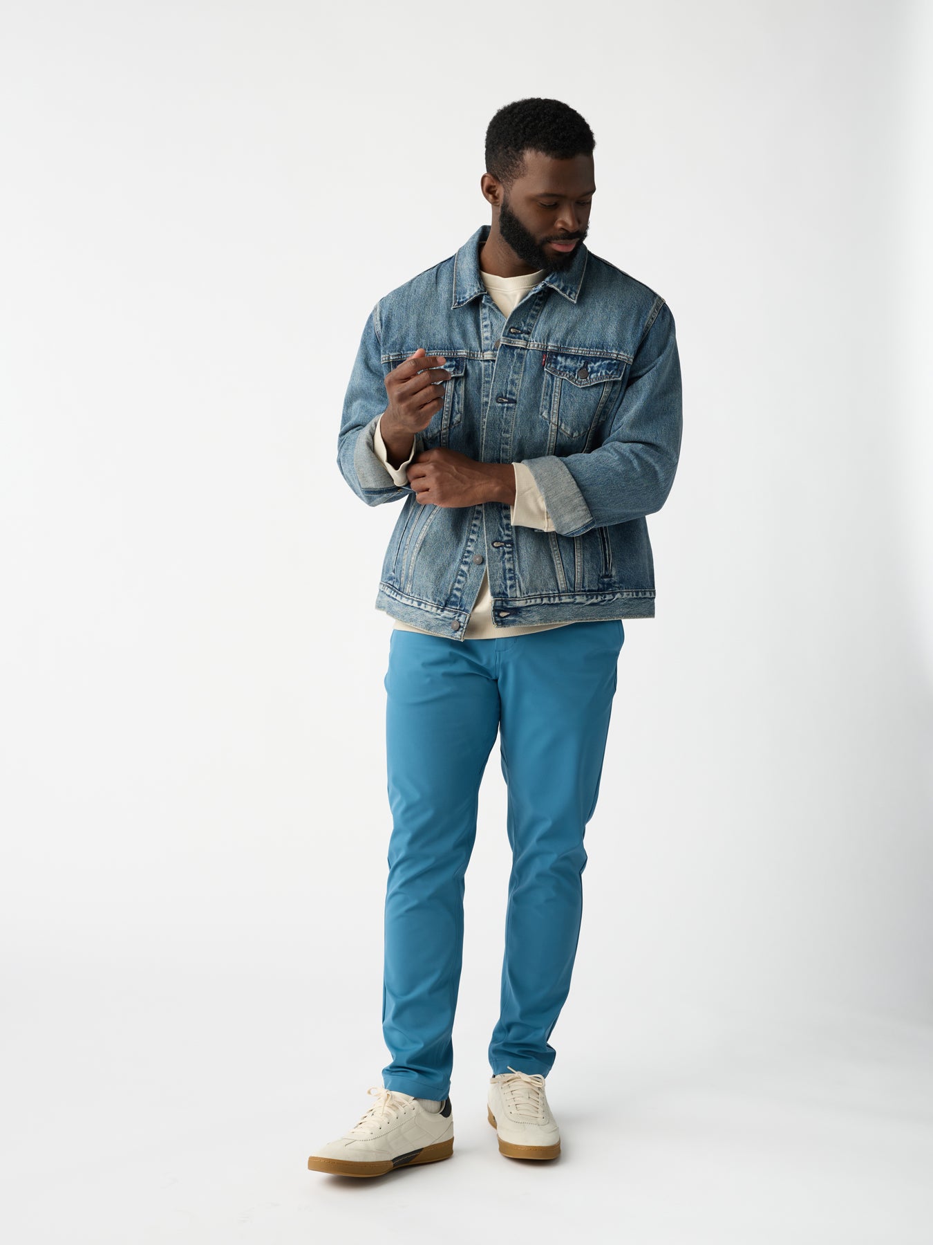 A man in Cozy Earth's Men's Everywhere Pant, paired with a denim jacket and light shirt, stands against a plain white background wearing white sneakers. He gazes down and slightly to the side. 