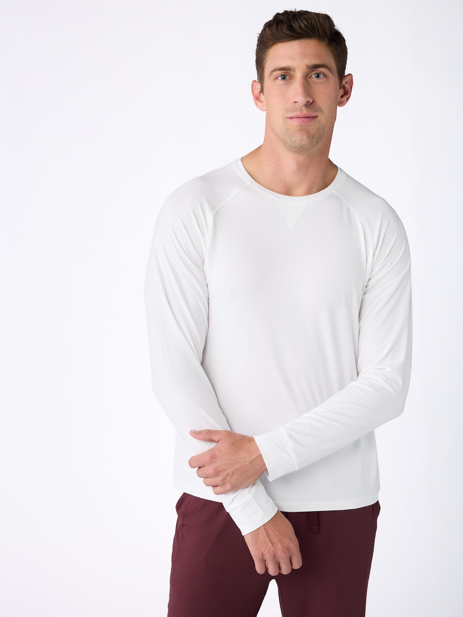 A man stands against a plain white background, dressed in the Men's Stretch-Knit Bamboo Long Sleeve from Cozy Earth and maroon pants. His arms are crossed casually, and he is gazing forward with a slight smile. 