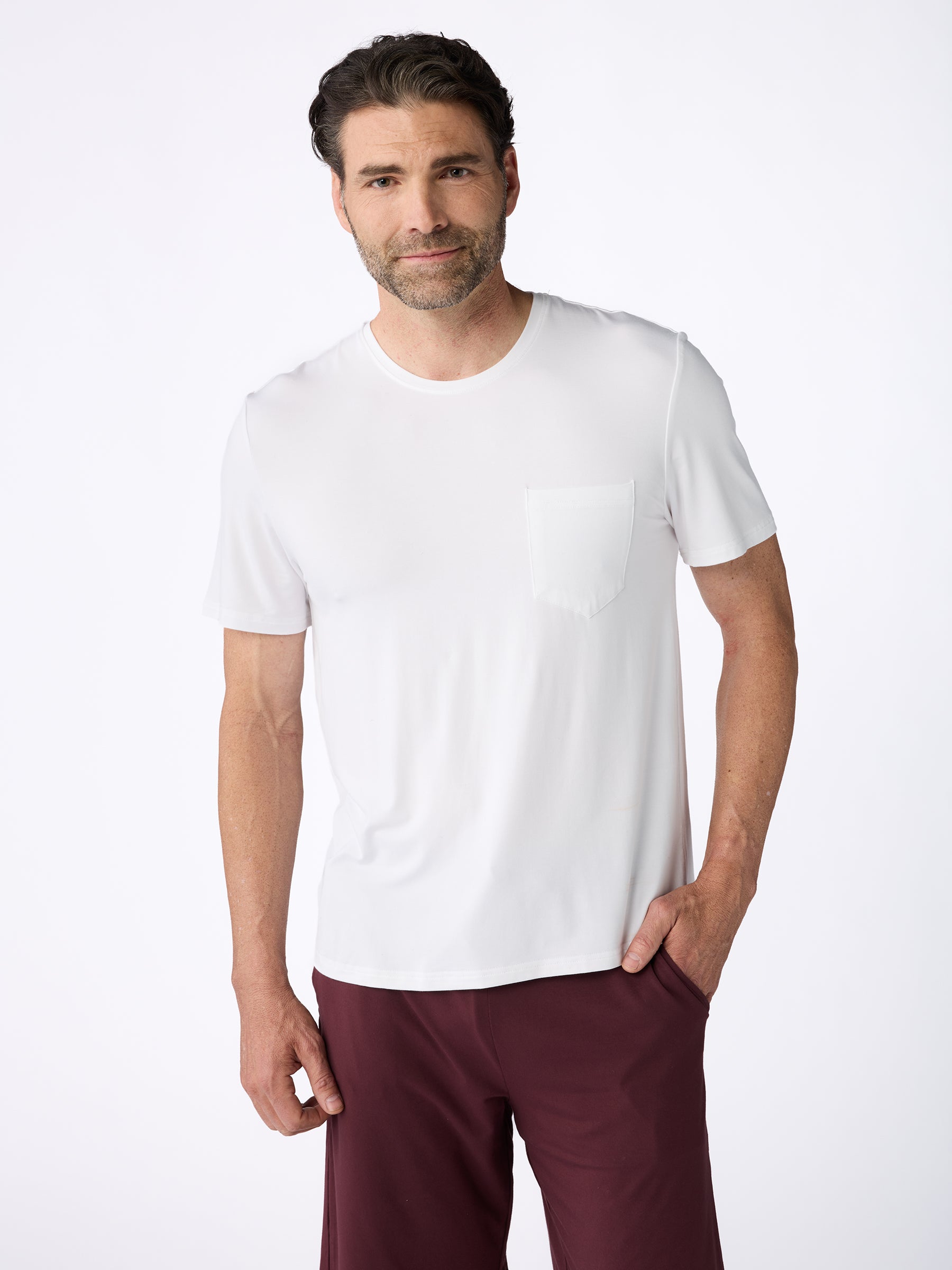 A man with short dark hair and a beard is wearing the Men's Stretch-Knit Bamboo Lounge Tee by Cozy Earth, paired with maroon pants. He is standing against a plain white background, looking at the camera with a relaxed expression. |Color:White