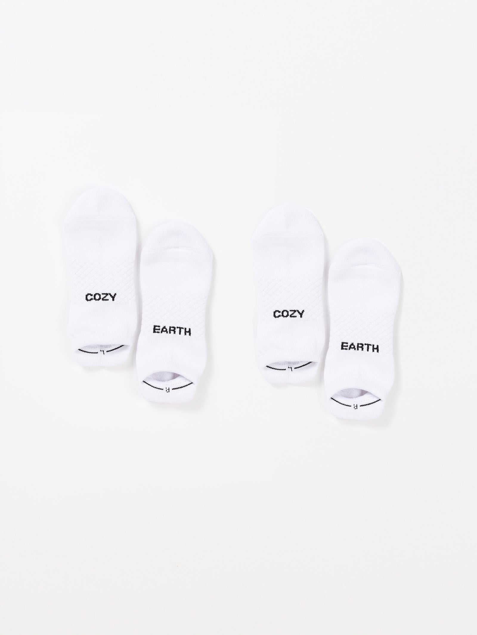 Two pairs of white Essential Ankle Socks from Cozy Earth are laid out on a plain background. The socks on the left have "COZY" written on the top and "EARTH" on the bottom. Similarly, the pair on the right features the same "COZY" and "EARTH" text. 