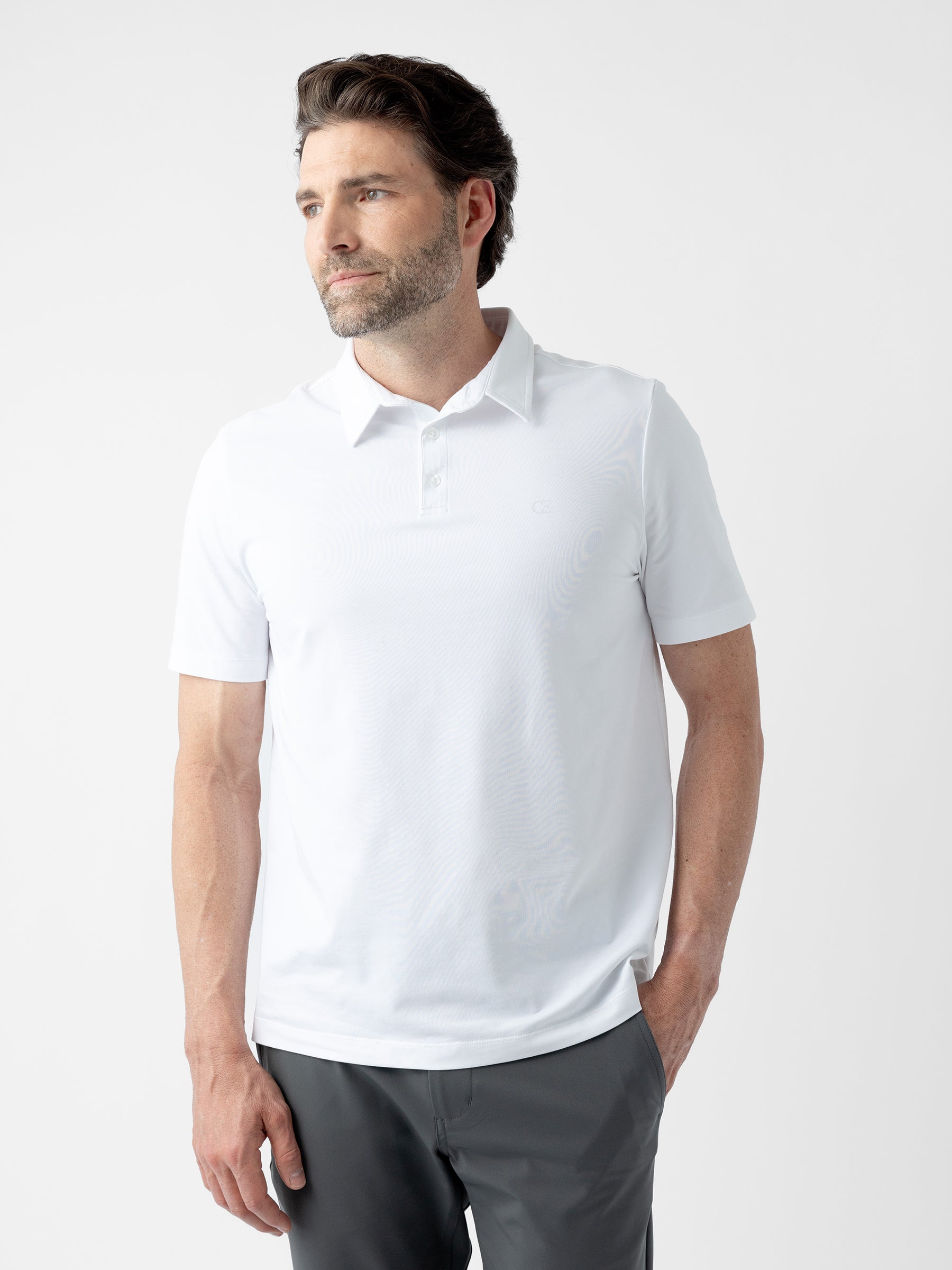 A man with short brown hair, a beard, and mustache is standing against a plain white background. He is wearing the Men's Everyday Polo from Cozy Earth in white and gray pants. One hand is in his pant pocket as he gazes to the side with a neutral expression. |Color:White