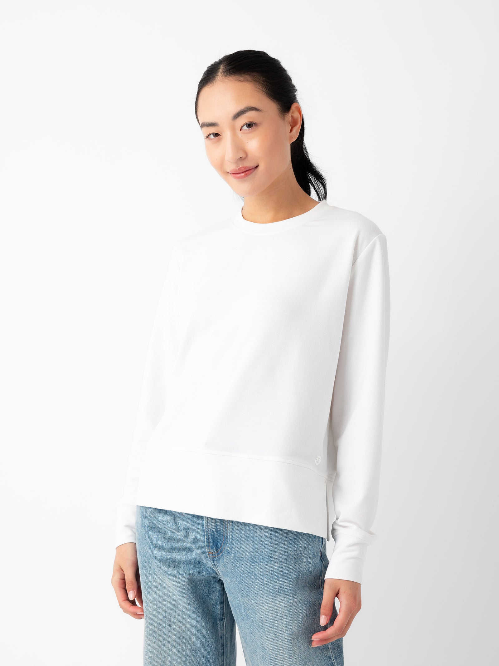 A person with long dark hair, wearing the Women's StudioLite Crewneck from Cozy Earth and light blue jeans, is standing against a plain white background. They are slightly smiling and have one hand in their pocket. 